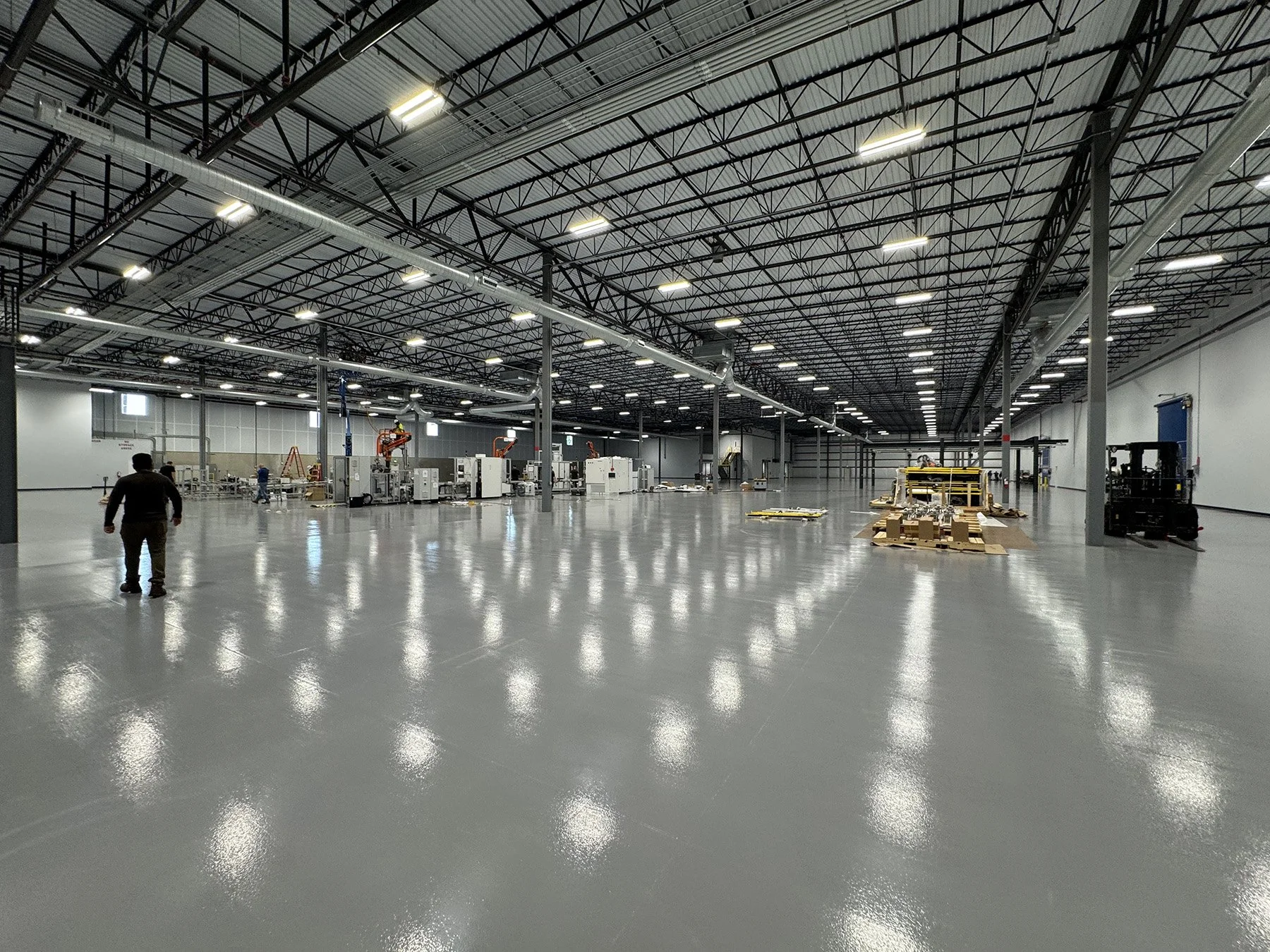 A large, empty industrial warehouse with a high ceiling, bright overhead lighting, and a smooth polished floor. There are a few workers and equipment scattered throughout, including machinery, pallets, and a forklift.