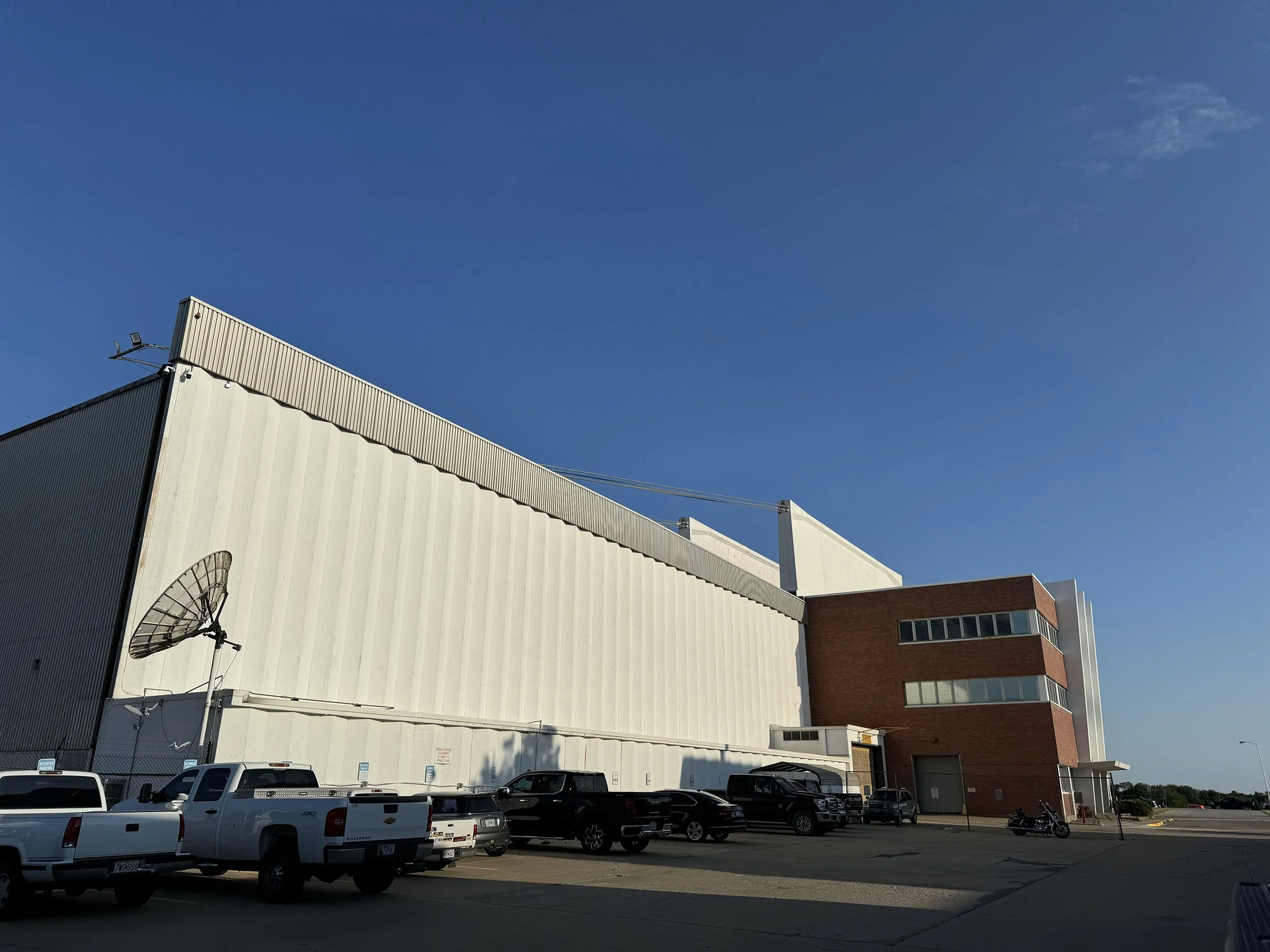 A large industrial building with cream-colored siding and a red section at the corner. Several cars and trucks are parked in a lot in front of the building. The sky above is clear and blue with a few wispy clouds.