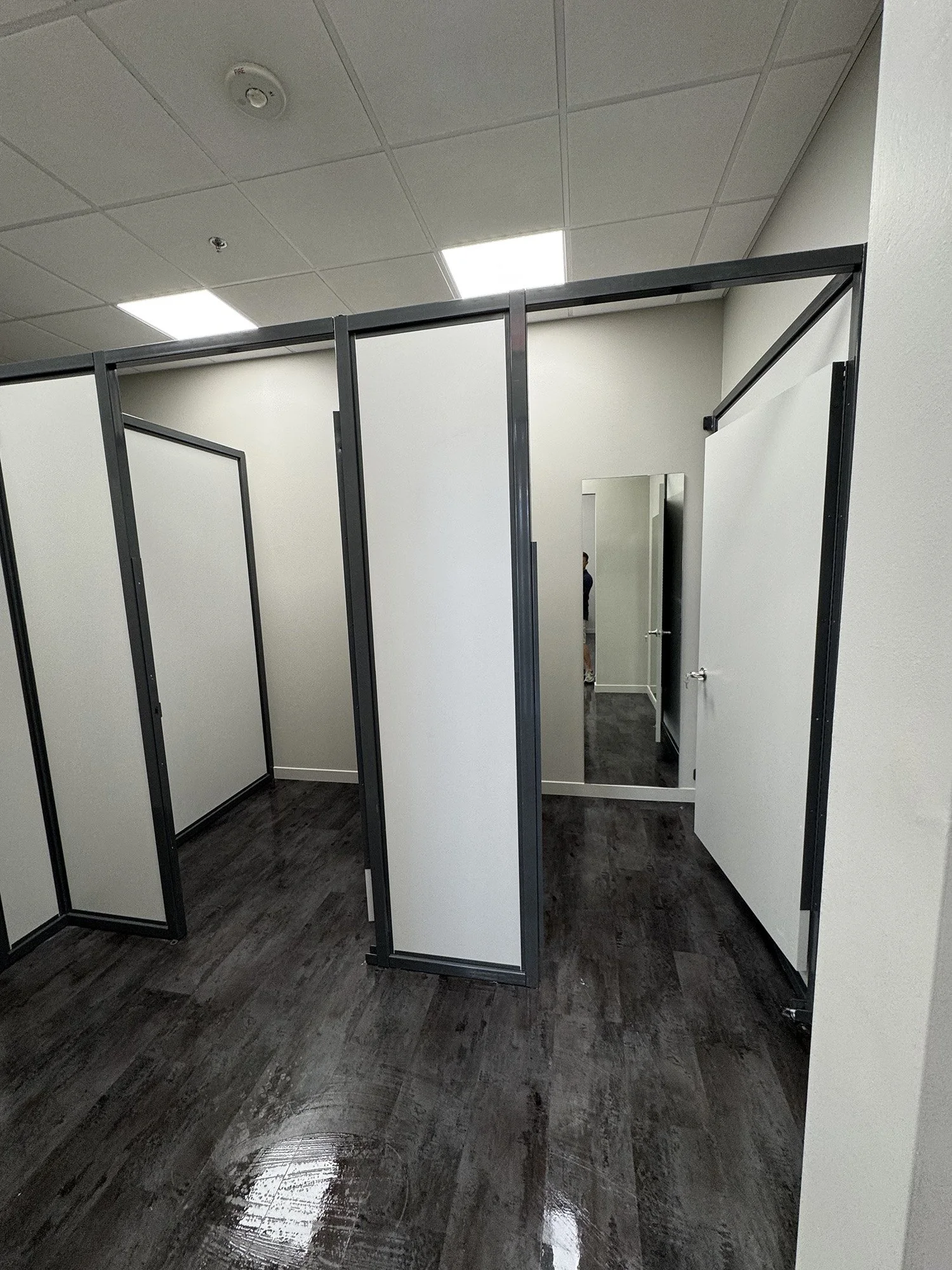Next generation toilet stall with white walls, black metal frame, and a mirror on the back wall, located in a public restroom with dark floor and ceiling lights.