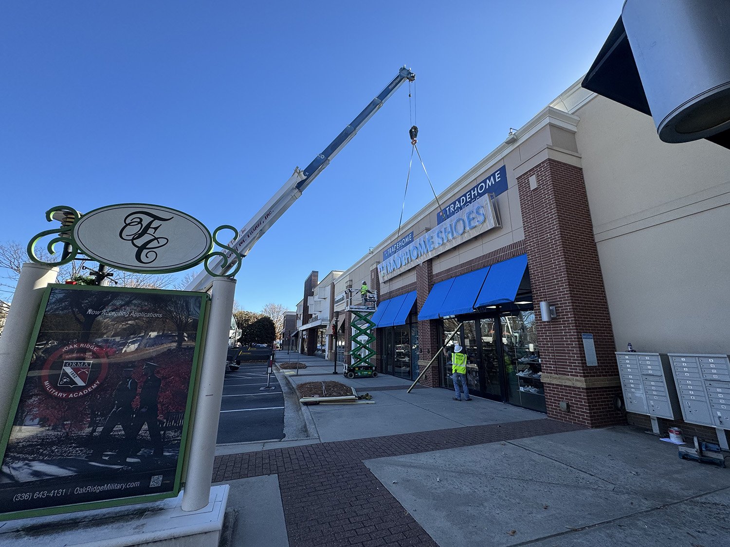 Construction workers installing blue signs and window canopies on a storefront labeled 'Tradehome.' Workers are elevated on a scissor lift, and the area is being prepared for opening.