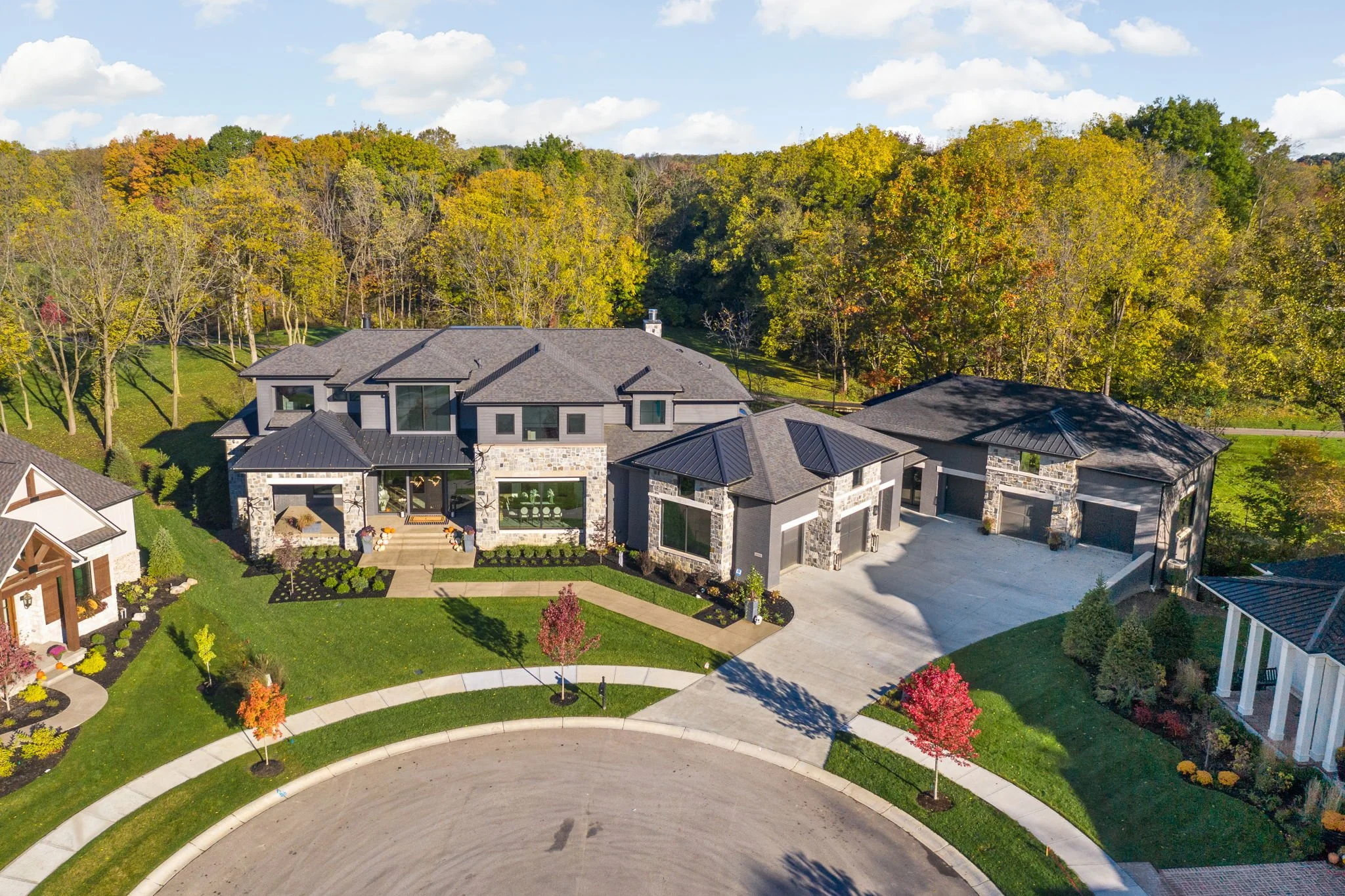 Aerial view of a modern, two-story house with a large driveway, surrounded by well-maintained lawns, trees, and autumn foliage. The house features a stone facade and multiple windows, set in a suburban neighborhood.