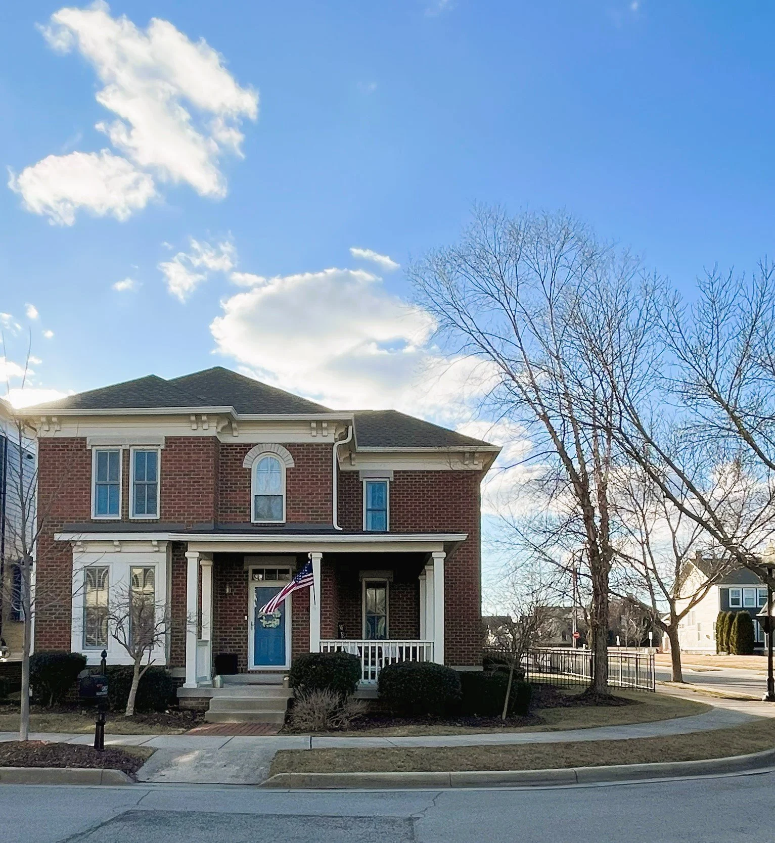 A two-story brick house with a blue front door, decorated with an American flag on the veranda. The house is surrounded by leafless trees and shrubs, with a clear blue sky in the background.