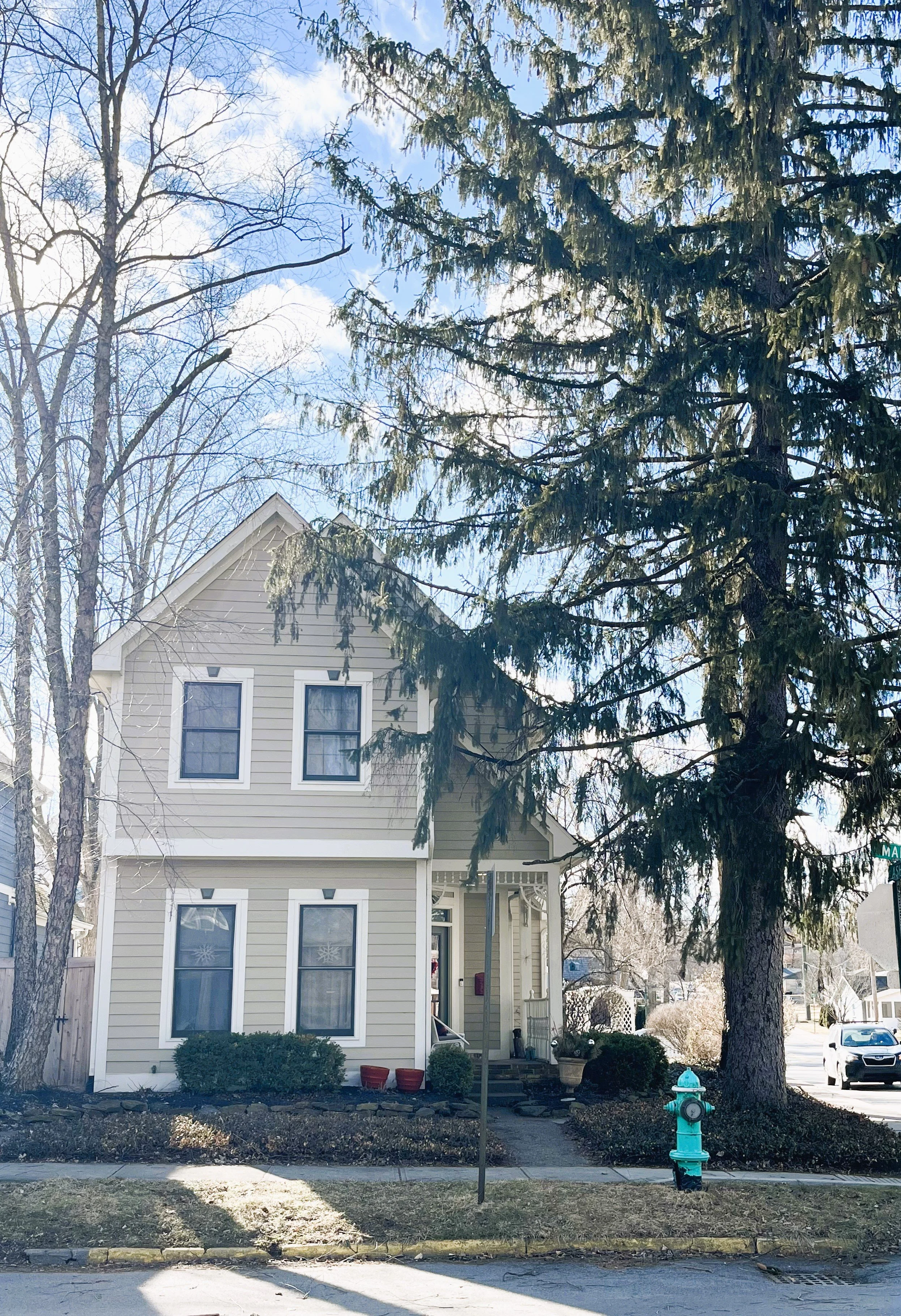 A beige two-story house with white trim, front porch, and large evergreen tree. There are bare trees and a turquoise fire hydrant on the sidewalk. The sky is clear with some clouds.