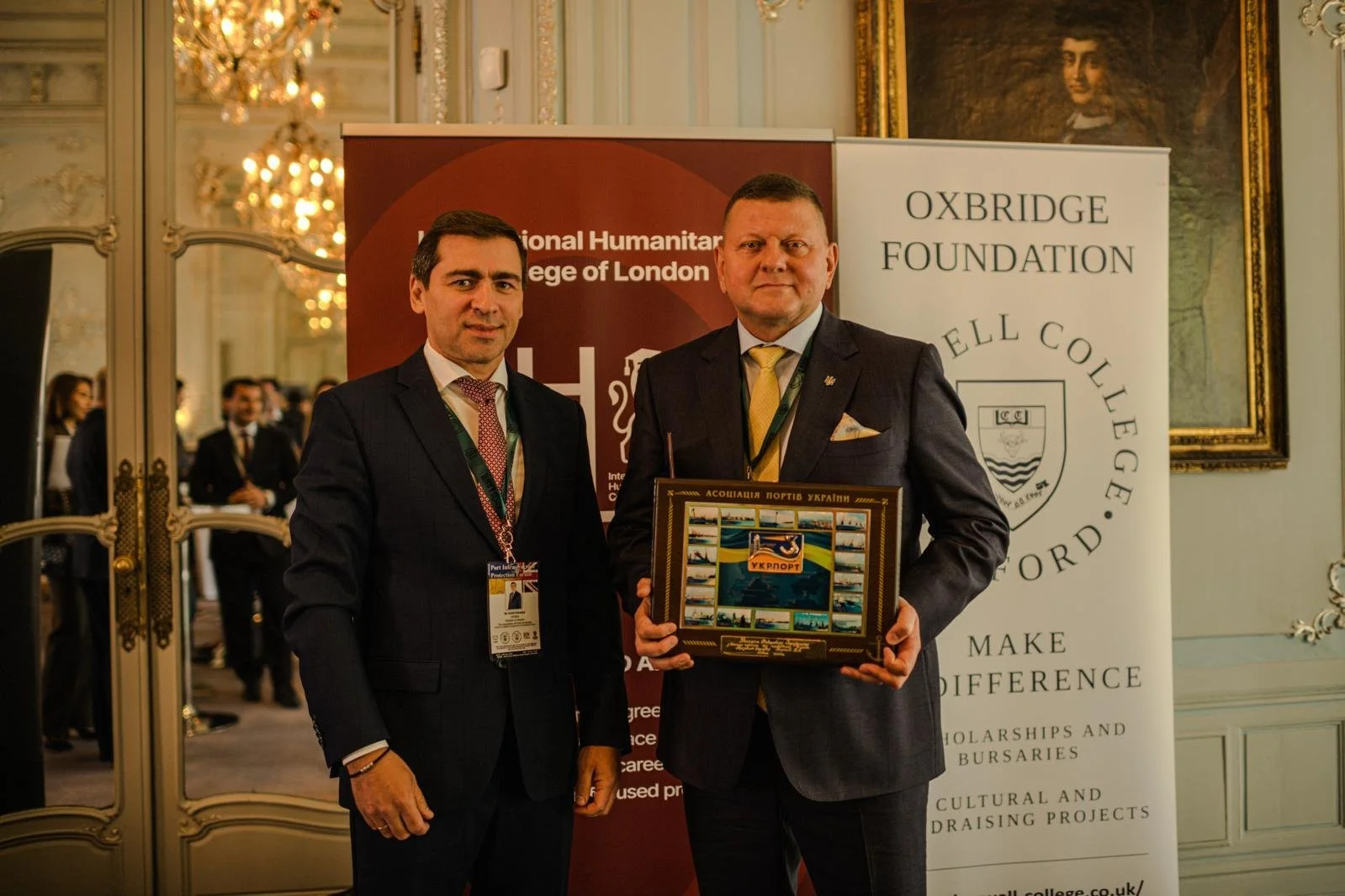 Two men in formal suits standing indoors in front of banners, one holding a framed collage of transportation images and Ukrainian symbols, at a formal event.