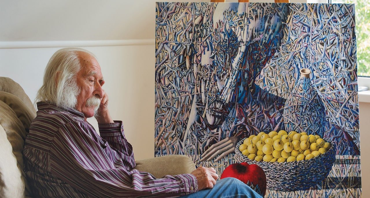 An elderly man with long white hair and a mustache sitting on a beige couch, talking on a cell phone, with an abstract painting of a person with a bicycle in the background. A large basket of yellow cherries and a red apple are on his lap.