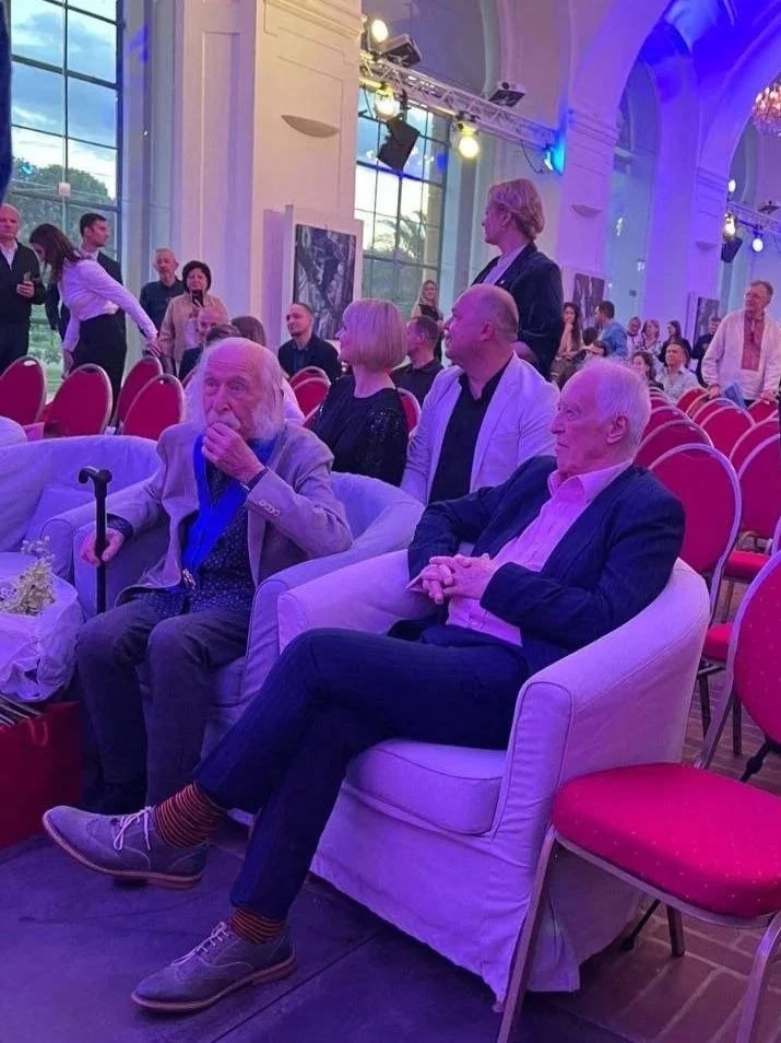 People seated in chairs and sofas at a formal event in a large hall with high arched ceiling and large windows. Some are talking and others are observing the event.