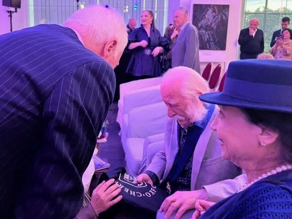 An elderly man with white hair and beard sits in a chair, looking at a book or item held by a woman with a blue hat and pearl necklace, who is smiling. A man with white hair and a woman with long hair in the background are also present, engaged in conversation. The setting appears to be indoors at a social event.