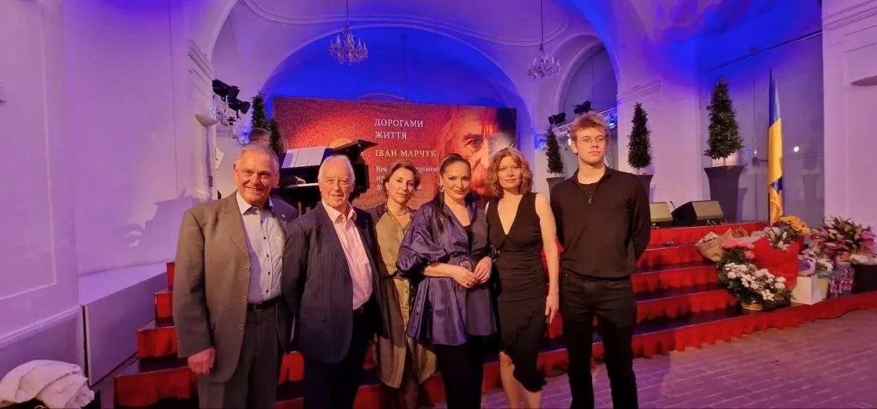 Six people standing on a stage with a red carpet, surrounded by flowers, with a large screen behind them displaying text and a portrait, and Ukrainian flag to the right.