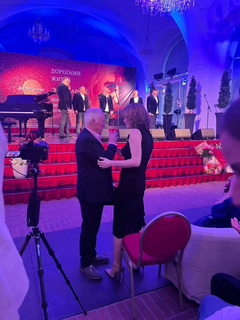 A man and a woman are dancing together at a formal event, with a stage and a band performing in the background. The event appears to be celebratory in nature, possibly an anniversary or tribute, with flowers and decorations adorning the scene.