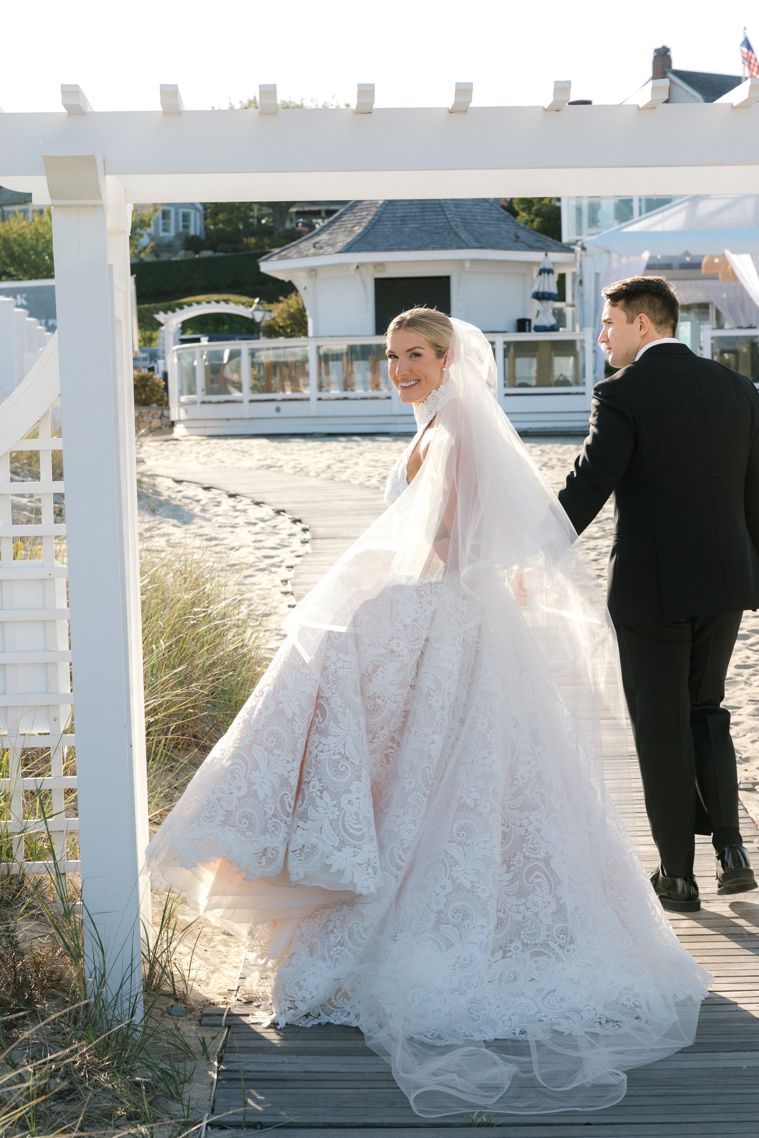 A bride in a lace wedding dress and veil holding hands with a groom in a black suit walking on a wooden boardwalk at a beachside venue.