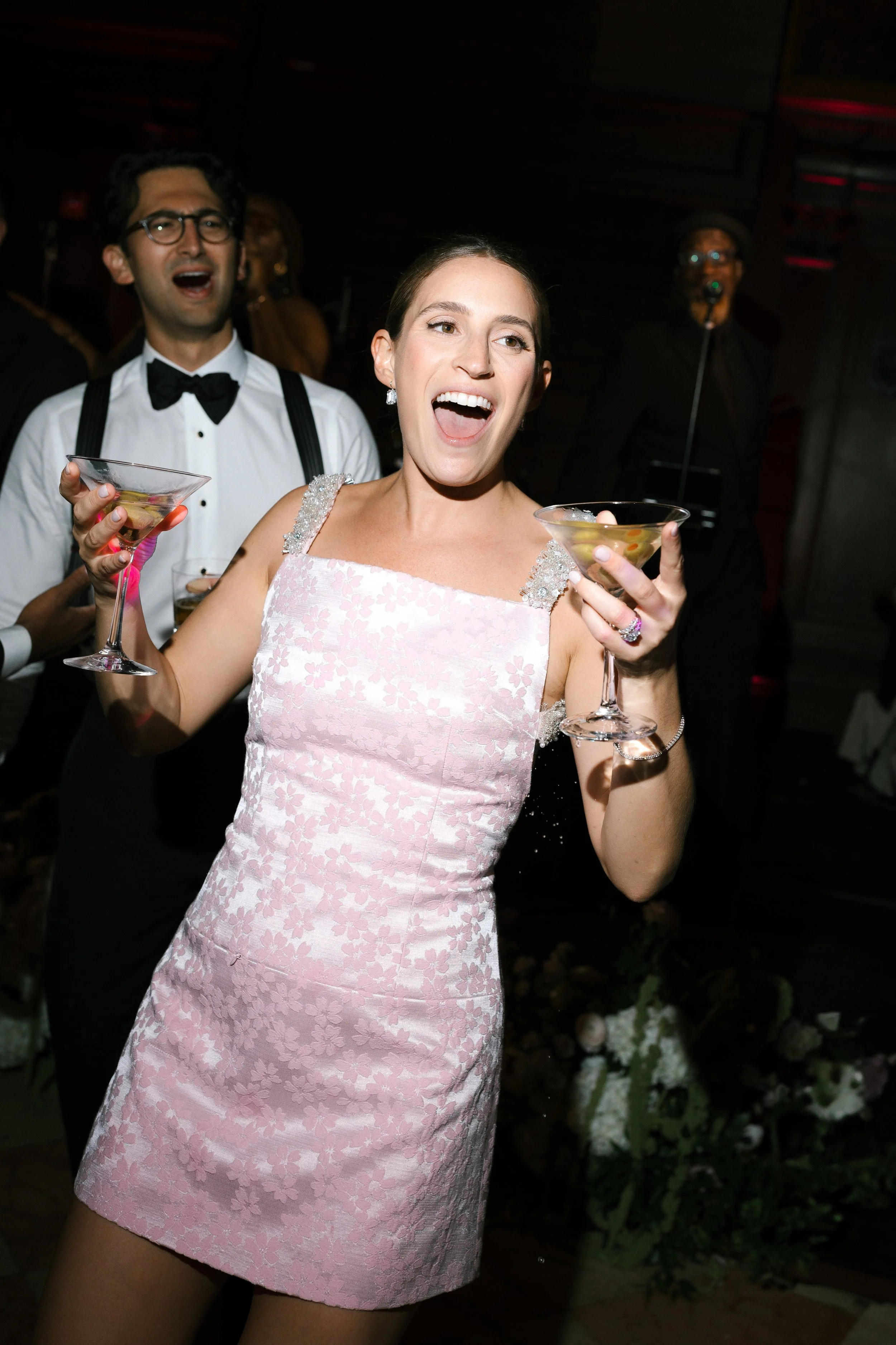 A woman in a pink floral dress celebrating with cocktails at a party, surrounded by other guests in formal attire.
