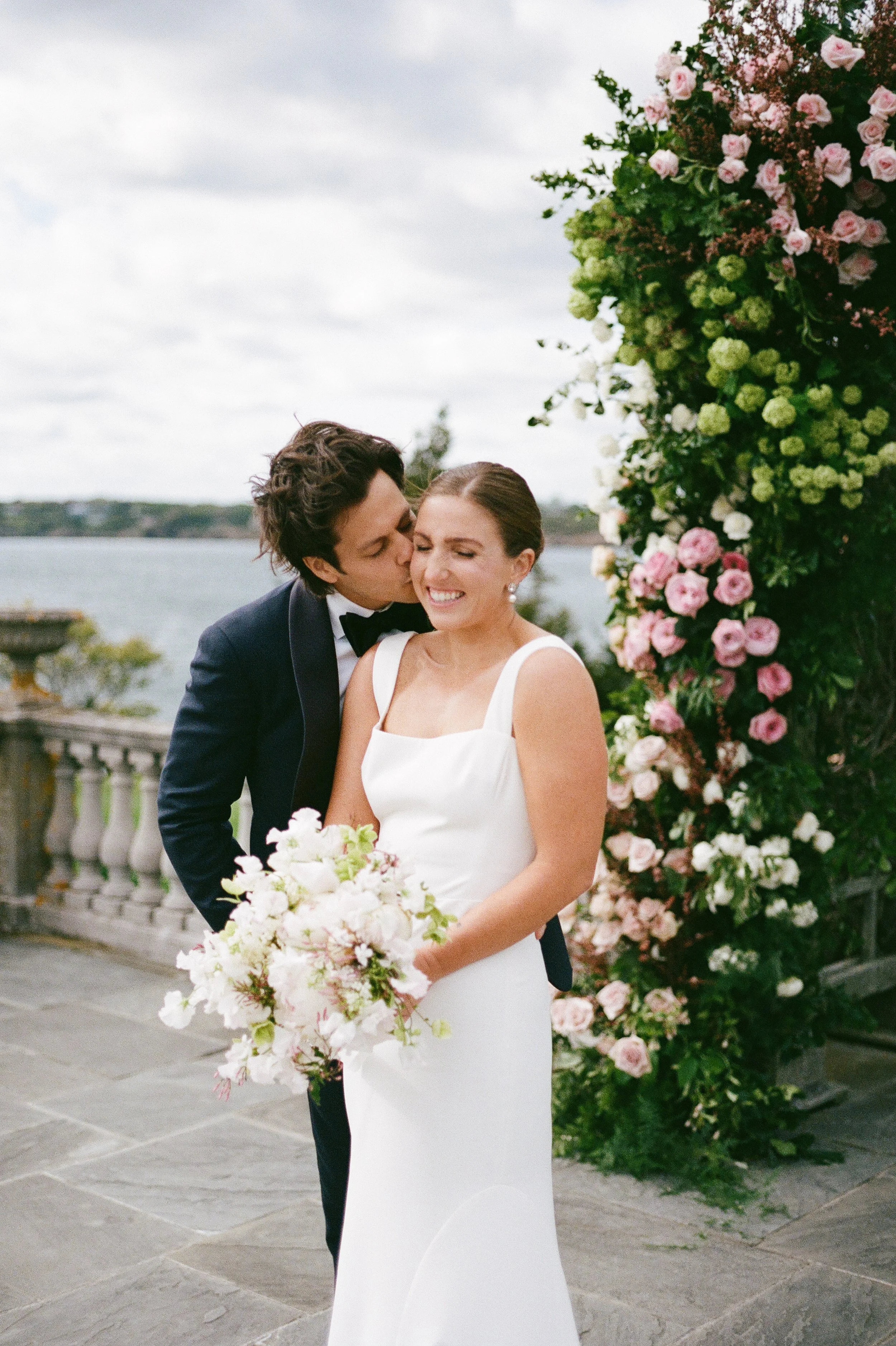 A bride in a white dress holding a bouquet, standing by a floral arch, as groom kisses her on the cheek outdoors with a body of water in the background.