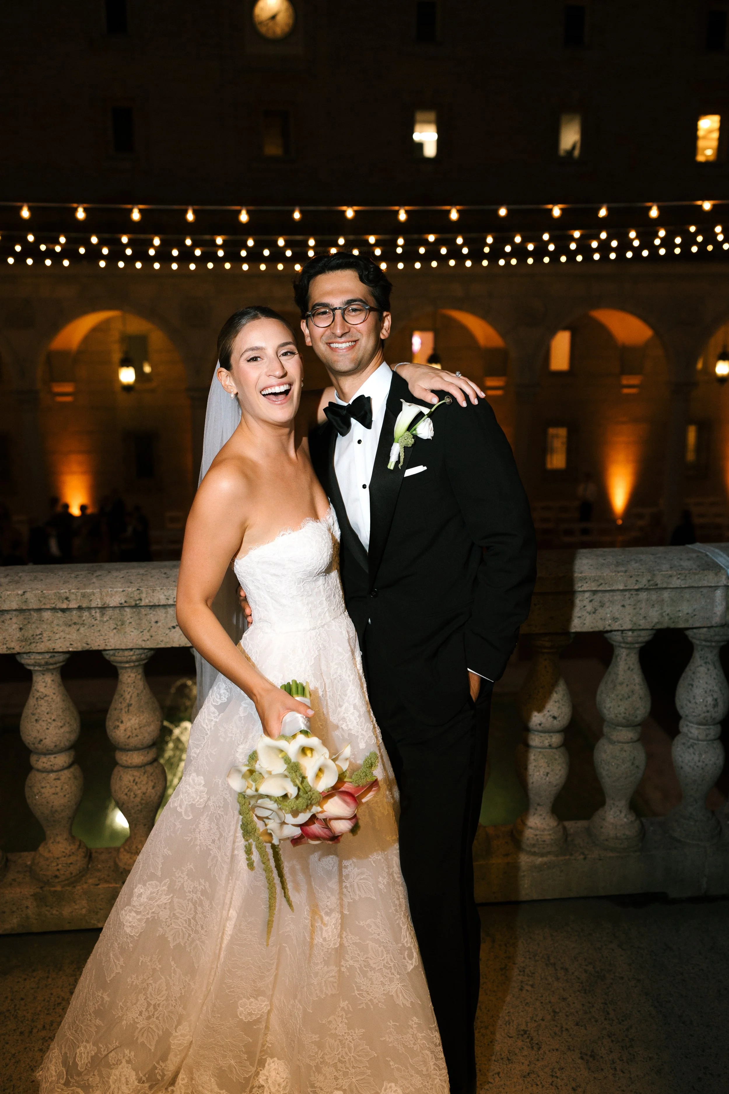 A newlywed couple standing on a balcony at night, smiling. The bride is holding a bouquet, wearing a strapless lace wedding gown and a veil. The groom is in a black tuxedo with a bow tie and a white boutonniere. String lights are hanging above them, 