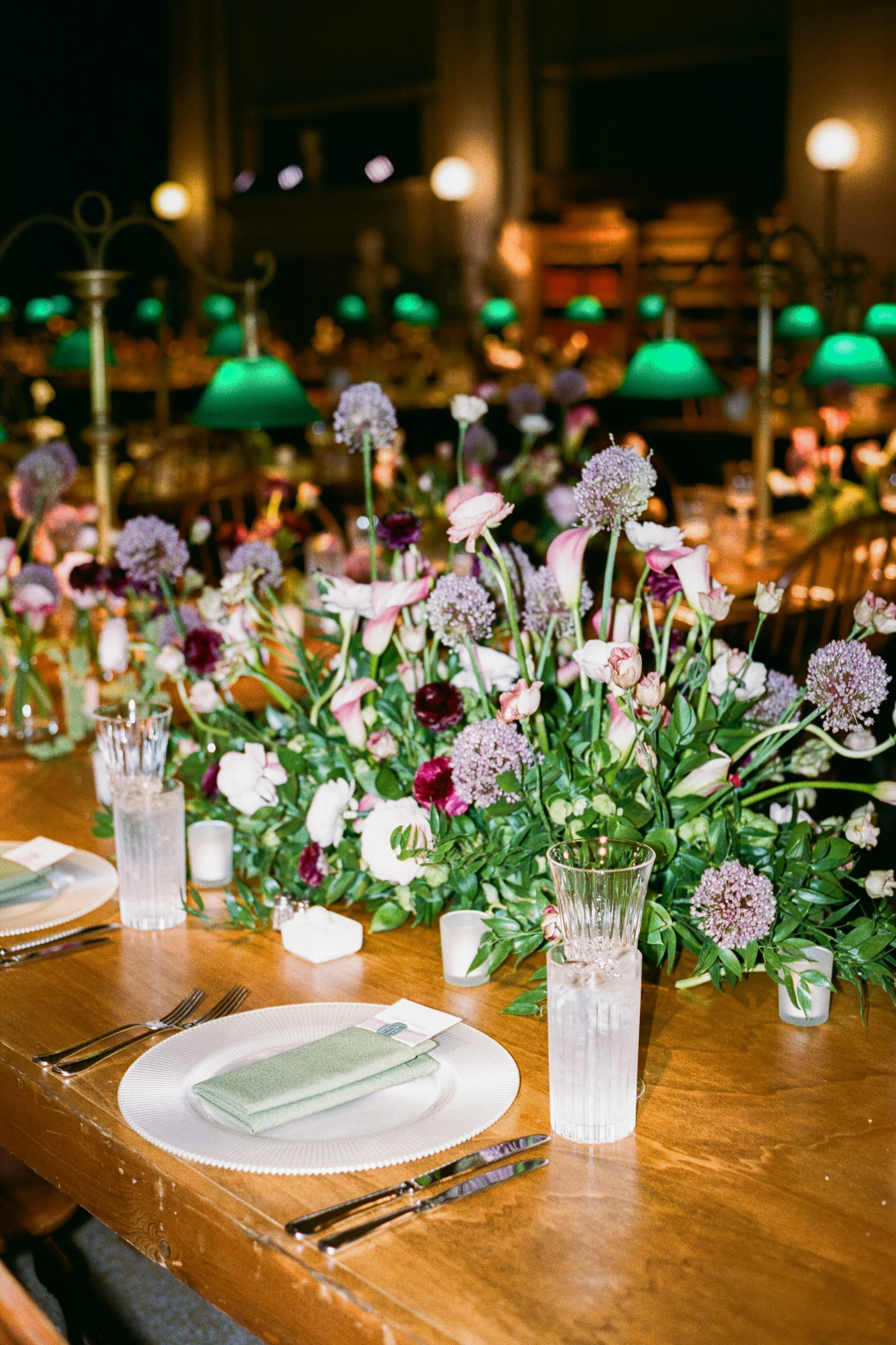 A decorated banquet table with a large floral centerpiece composed of purple, pink, and white flowers, set with clear glass tumblers, white plates, silver forks and knives, light green napkins, and small candles. The background shows soft, warm light