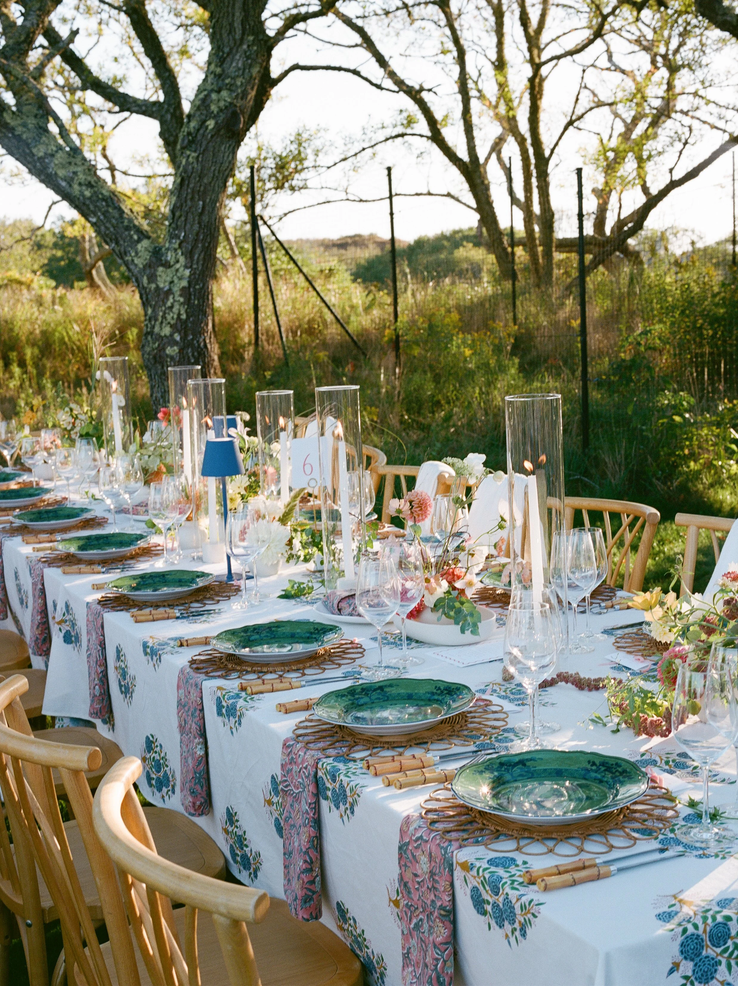 A long outdoor dining table set with plates, wine glasses, floral centerpieces, and bamboo placemats, surrounded by wooden chairs, with trees and greenery in the background.