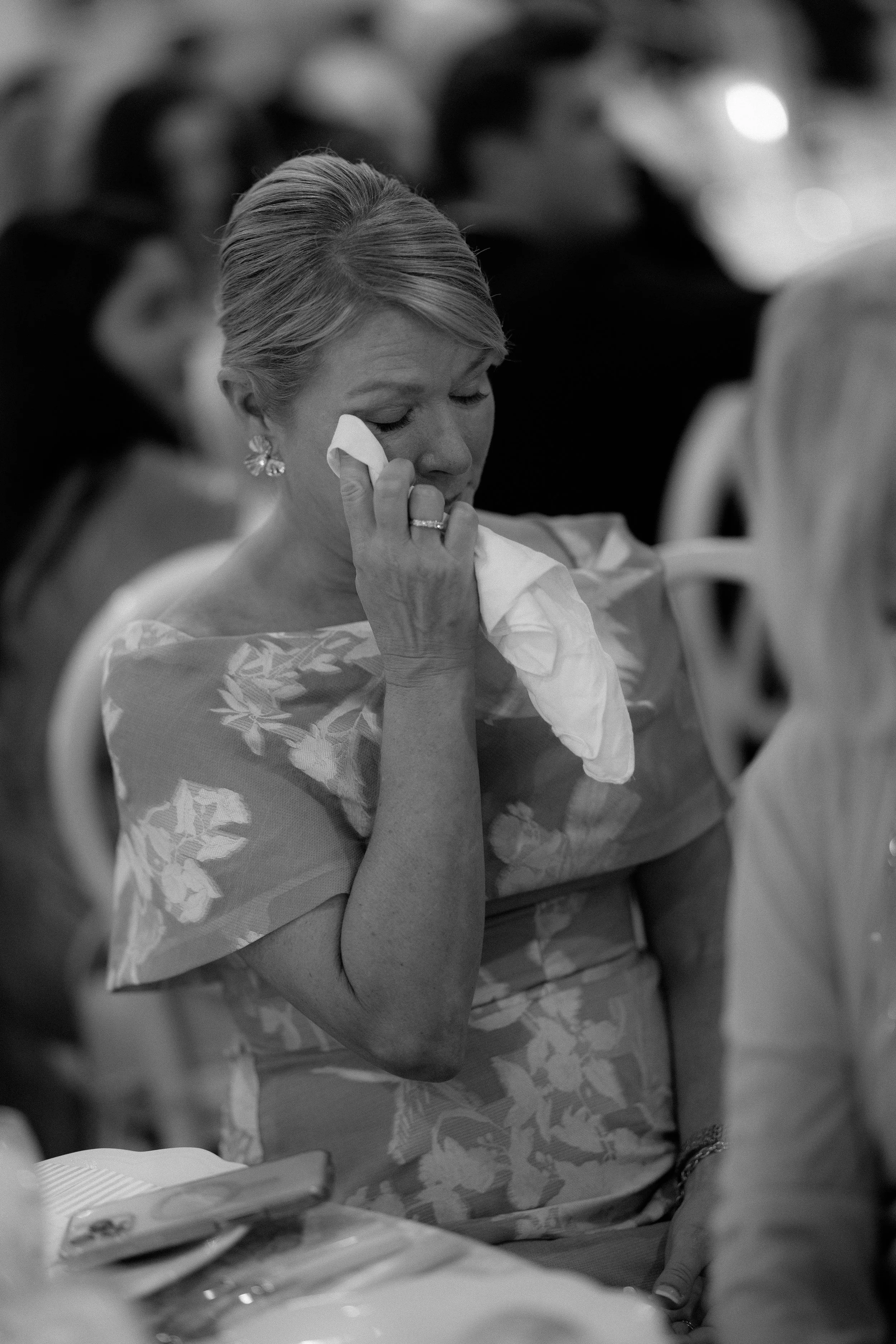 A woman in a floral dress is wiping tears from her eyes with a tissue during an emotional moment at a formal gathering.