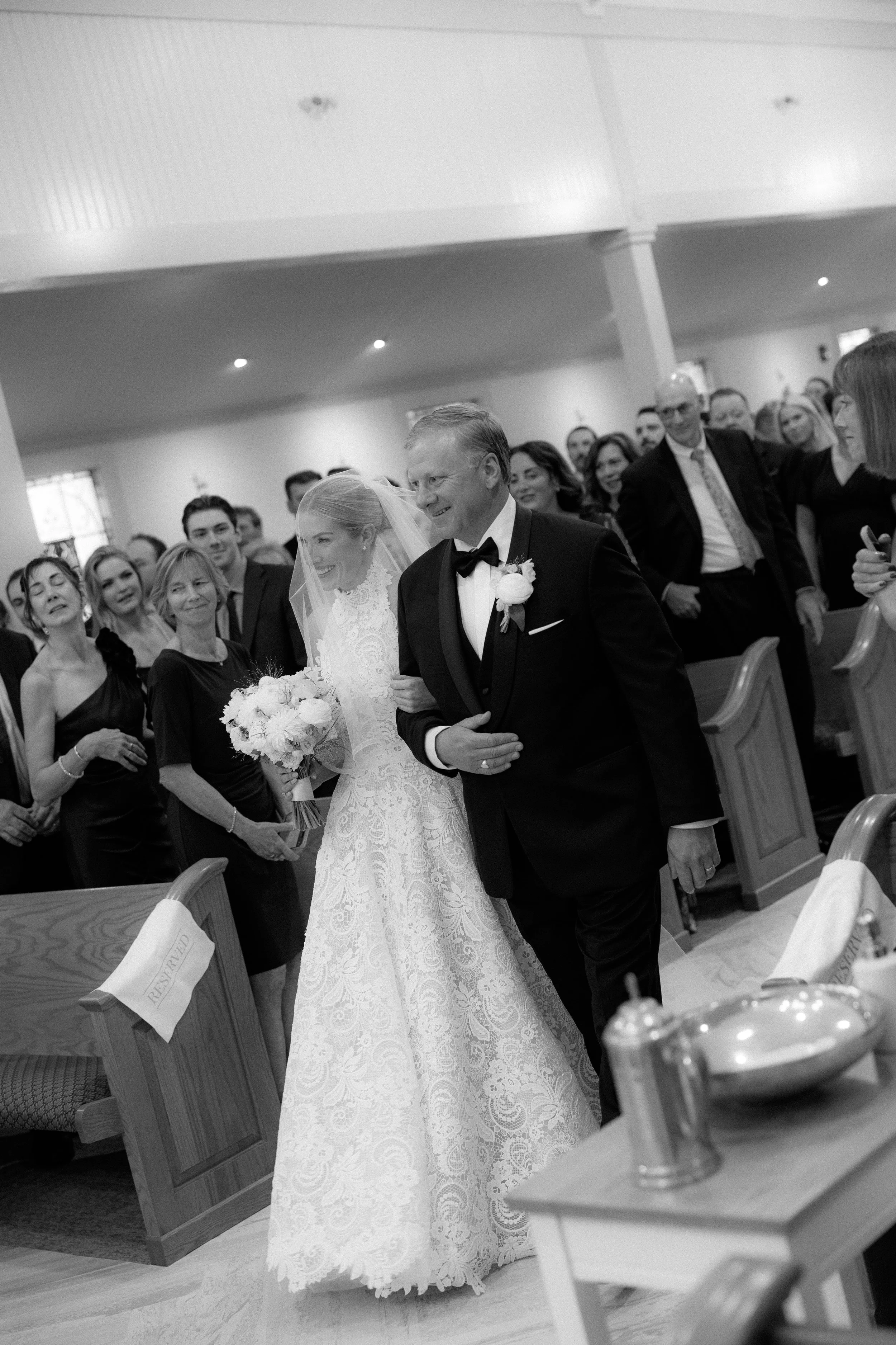 A bride walking down the aisle with her father at a wedding ceremony, surrounded by guests in a church or chapel.