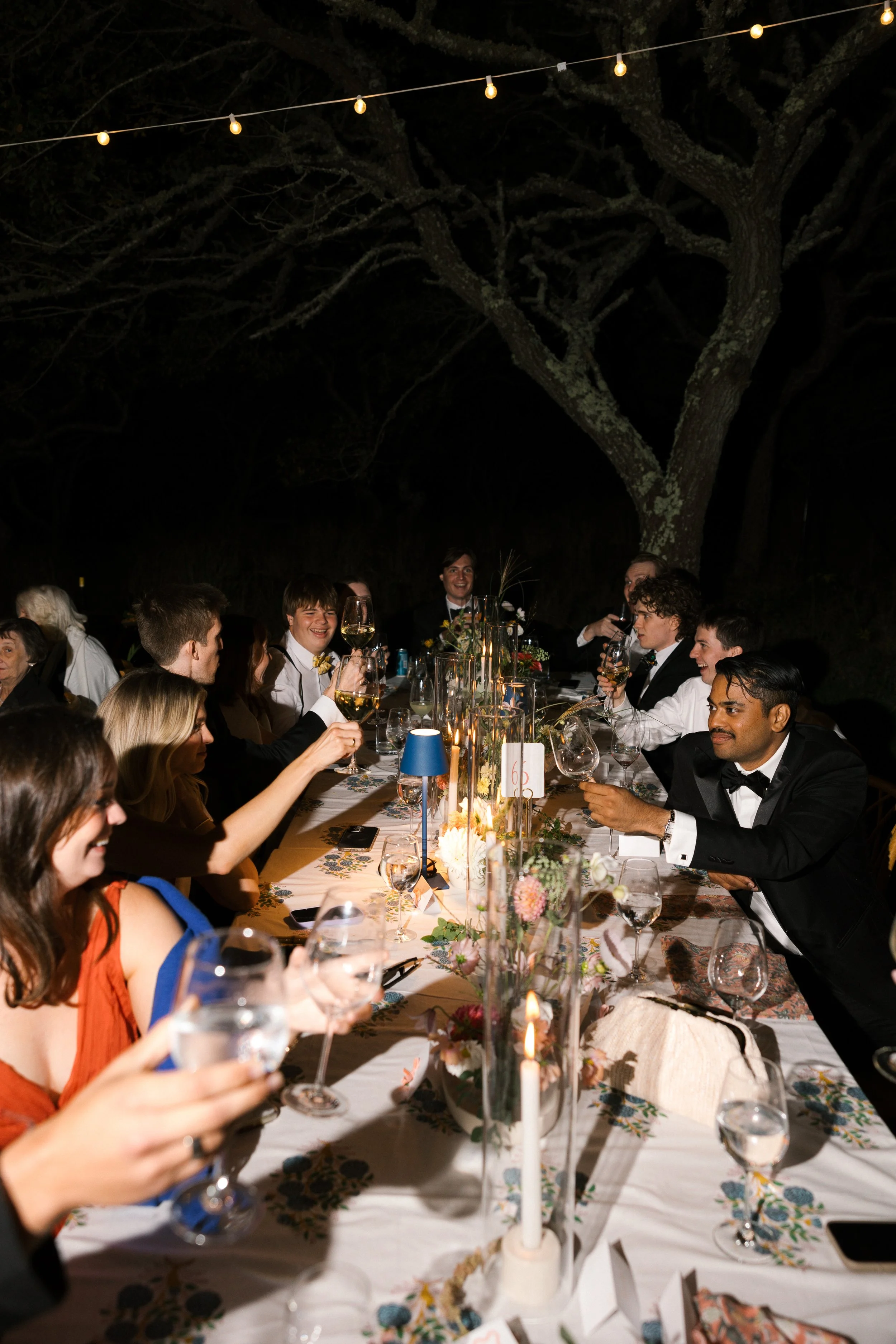 People celebrating at a long dinner table outdoors at night, with string lights overhead, floral centerpieces, and candles.