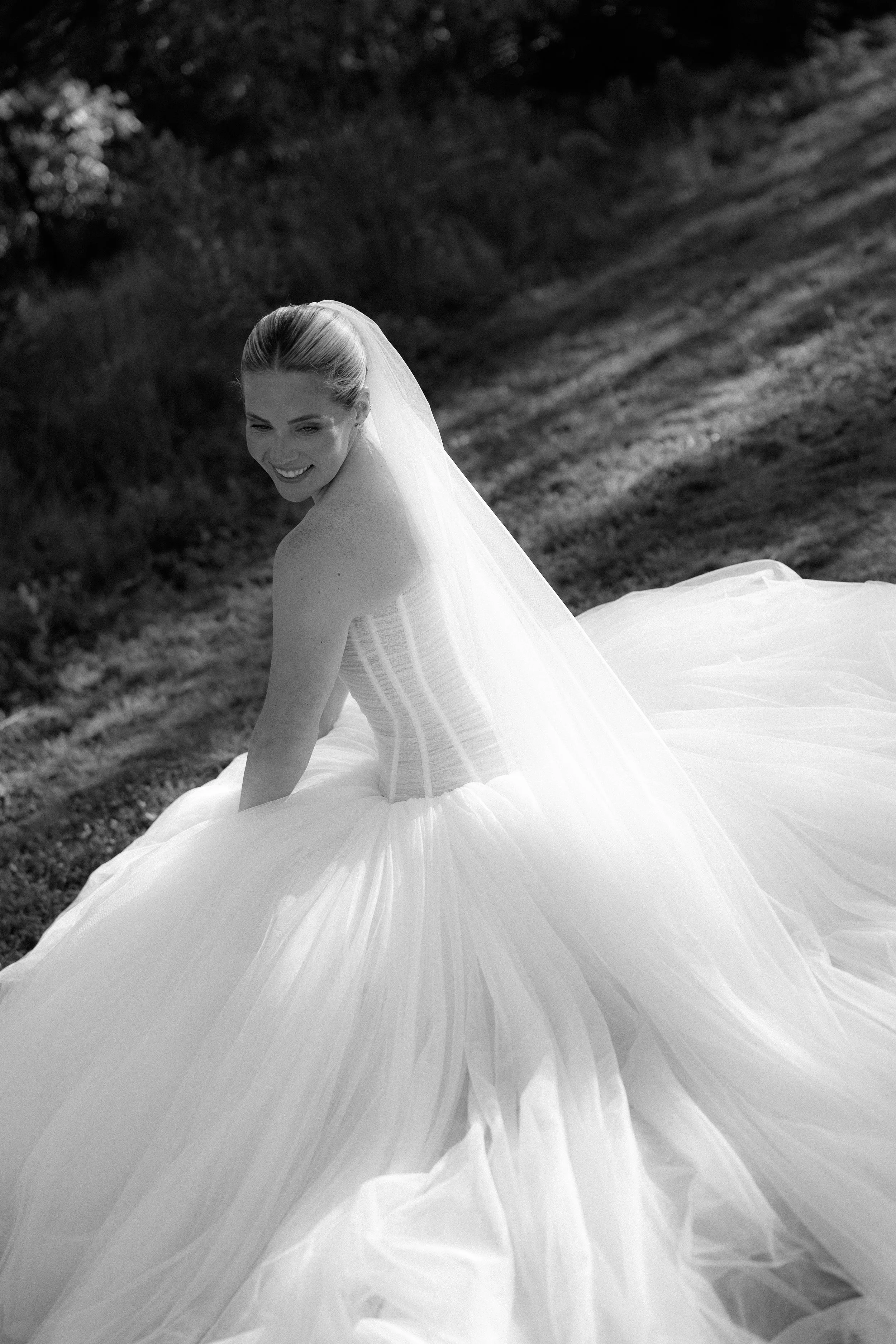 Black and white photo of a smiling bride in a strapless wedding gown with a full, layered skirt and veil, standing outdoors with a grassy background.