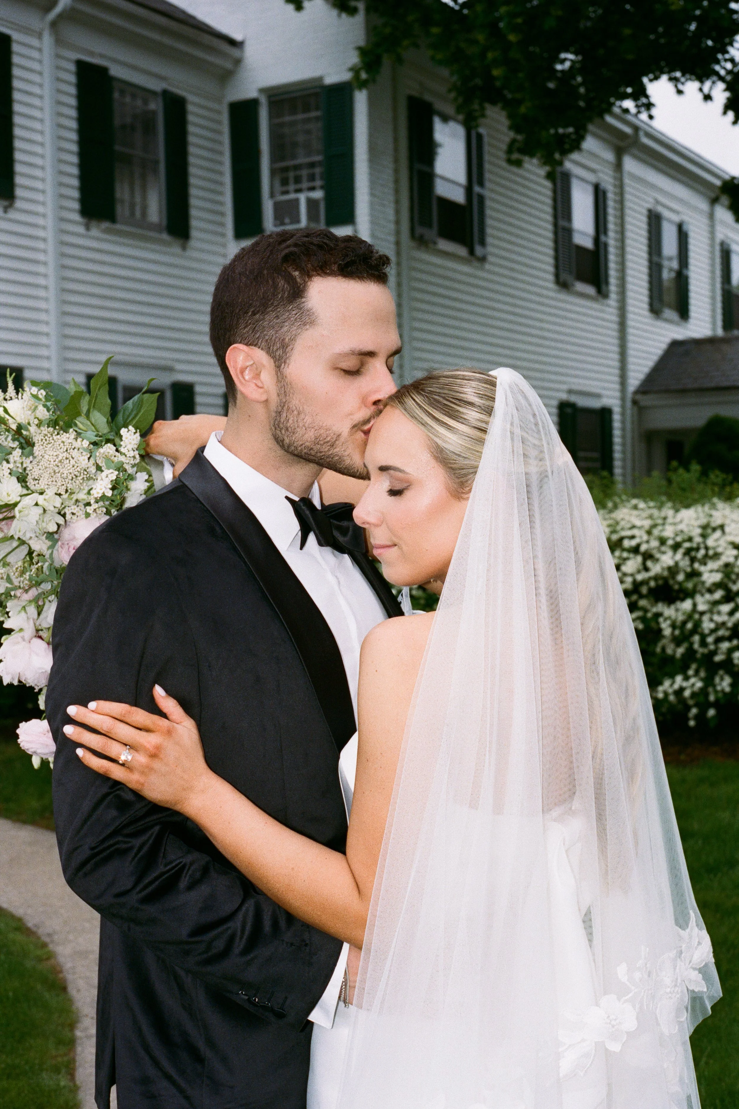A bride and groom embracing outside a house, with the groom kissing the bride's forehead. The groom is wearing a tuxedo and the bride a wedding dress with a veil.