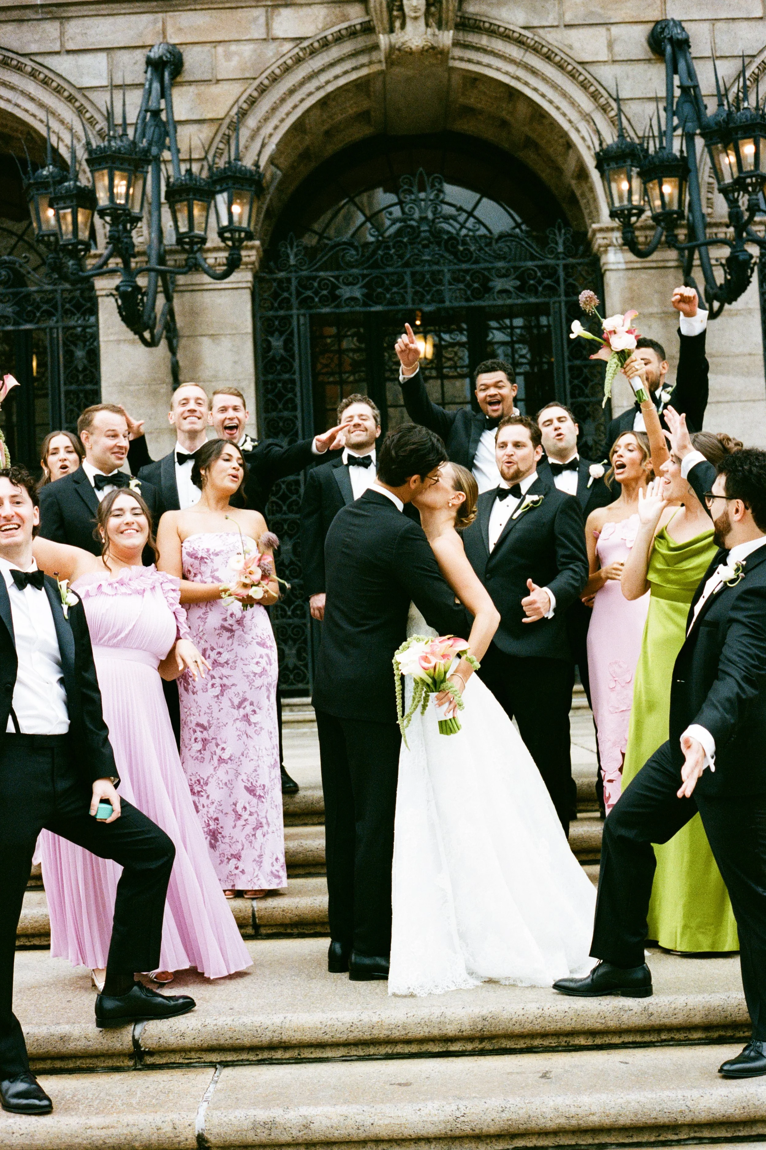 A wedding party celebrating outside a historic building, with the bride and groom kissing in front. The group includes bridesmaids and groomsmen, dressed in formal attire, some holding flowers, cheering and raising their hands.