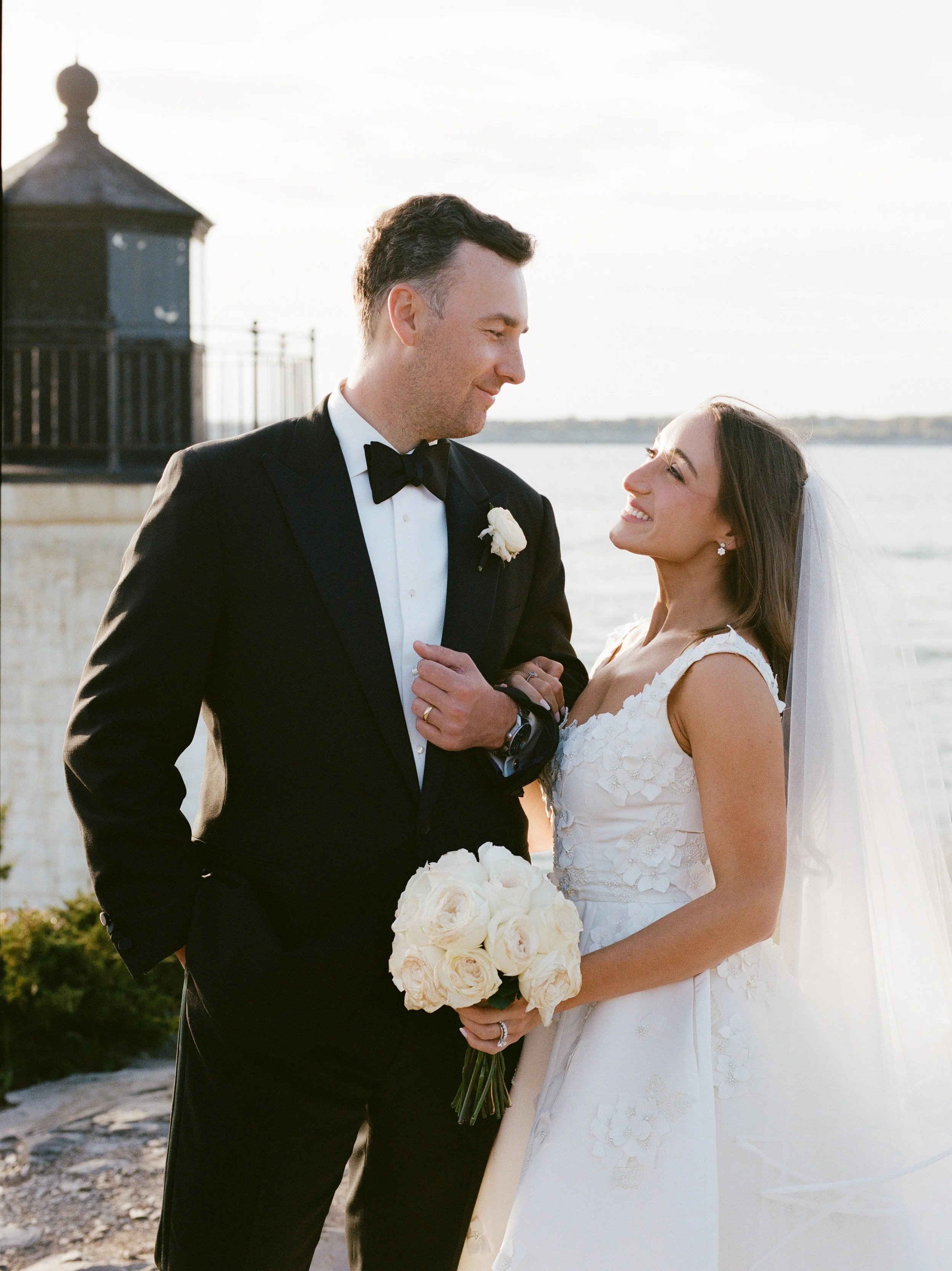 A newlywed couple gazes at each other, with the groom in a black tuxedo and the bride in a white wedding dress, holding a bouquet of white roses near a body of water during sunset.