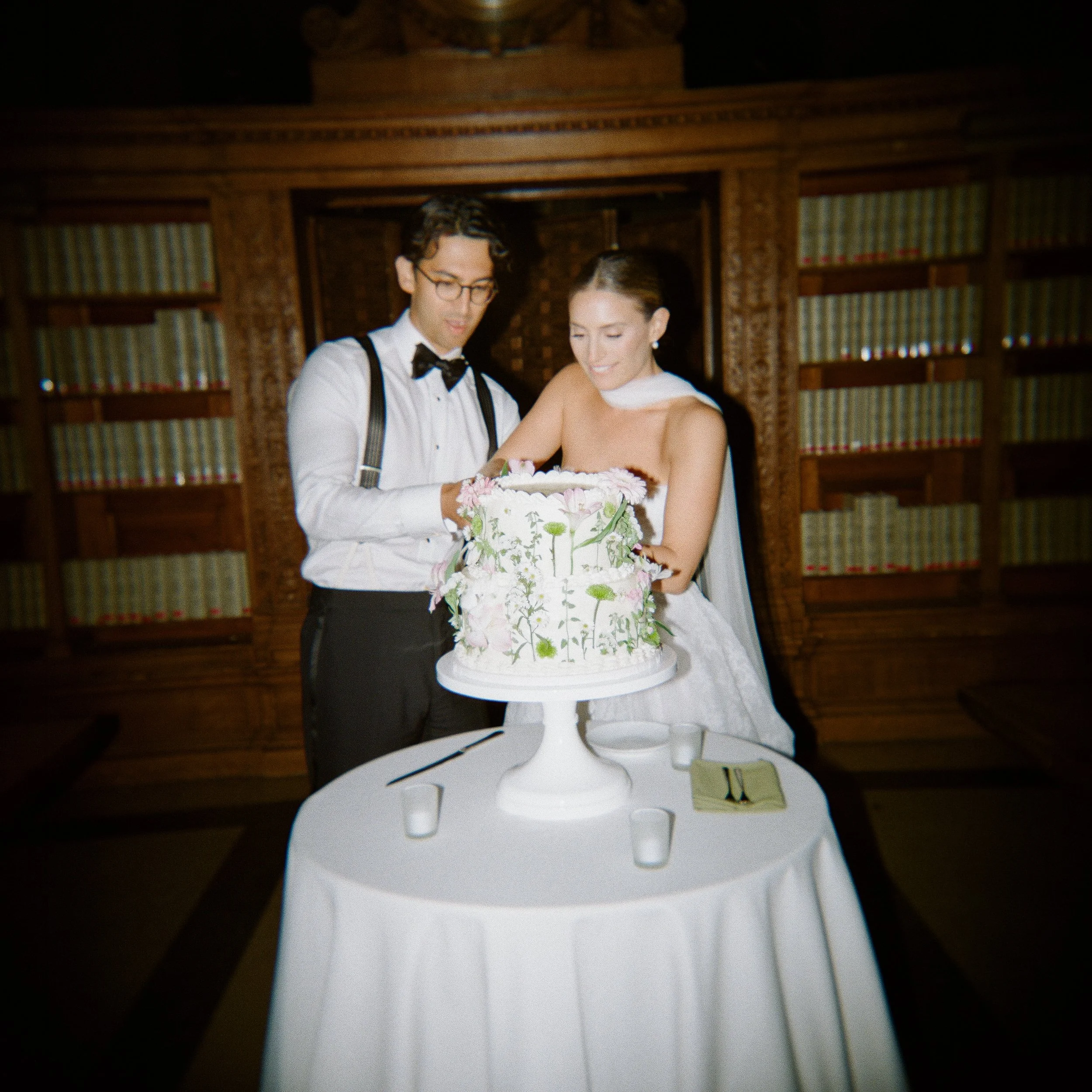 A bride and groom cutting a wedding cake together at their wedding reception, standing behind a white table with the cake on a white cake stand, decorated with white and pink flowers. The background features wooden bookshelves filled with books.