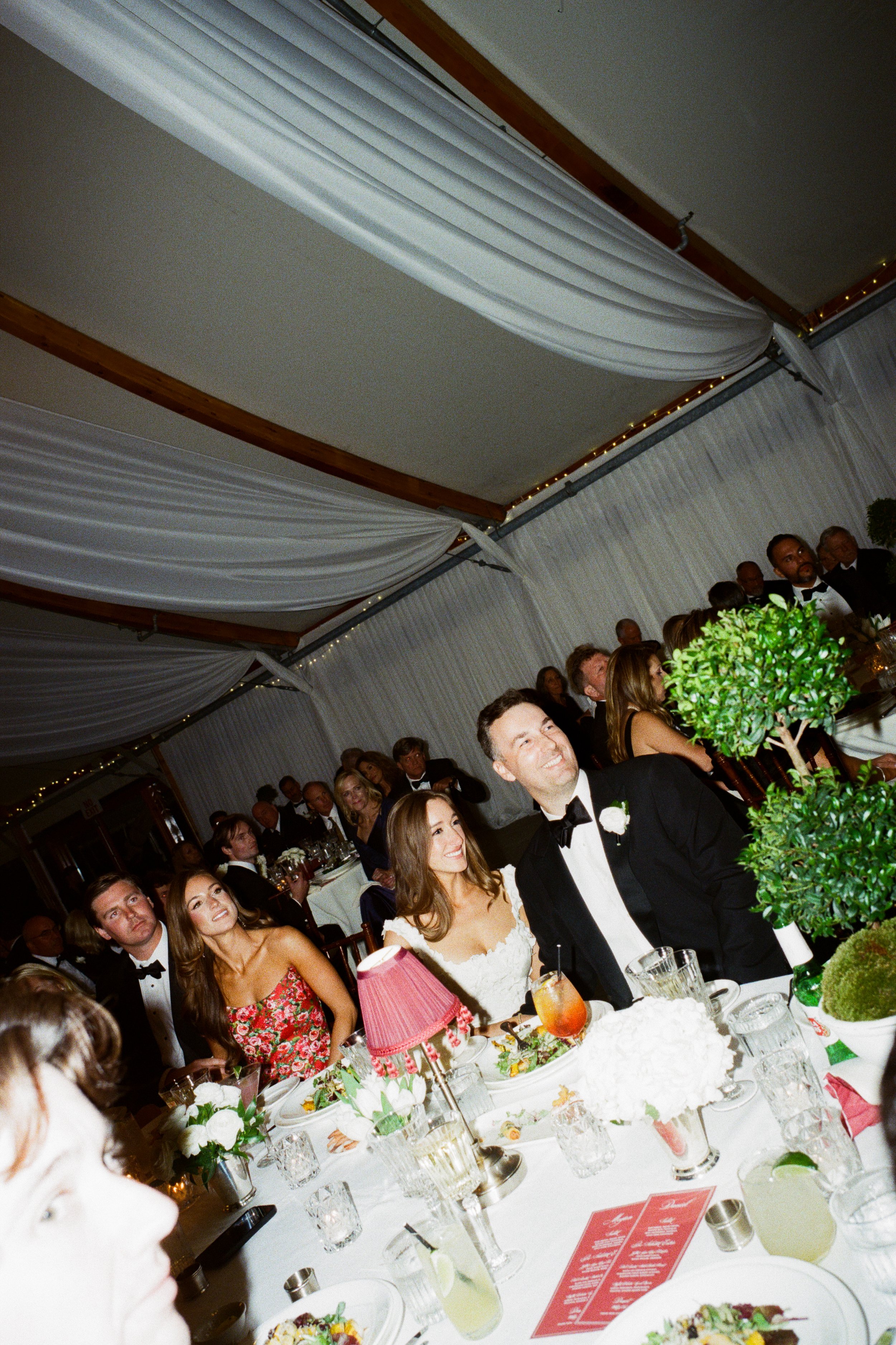 Group of people at a wedding reception, seated at decorated tables, with a bride and groom sitting together, smiling, surrounded by guests in formal attire.