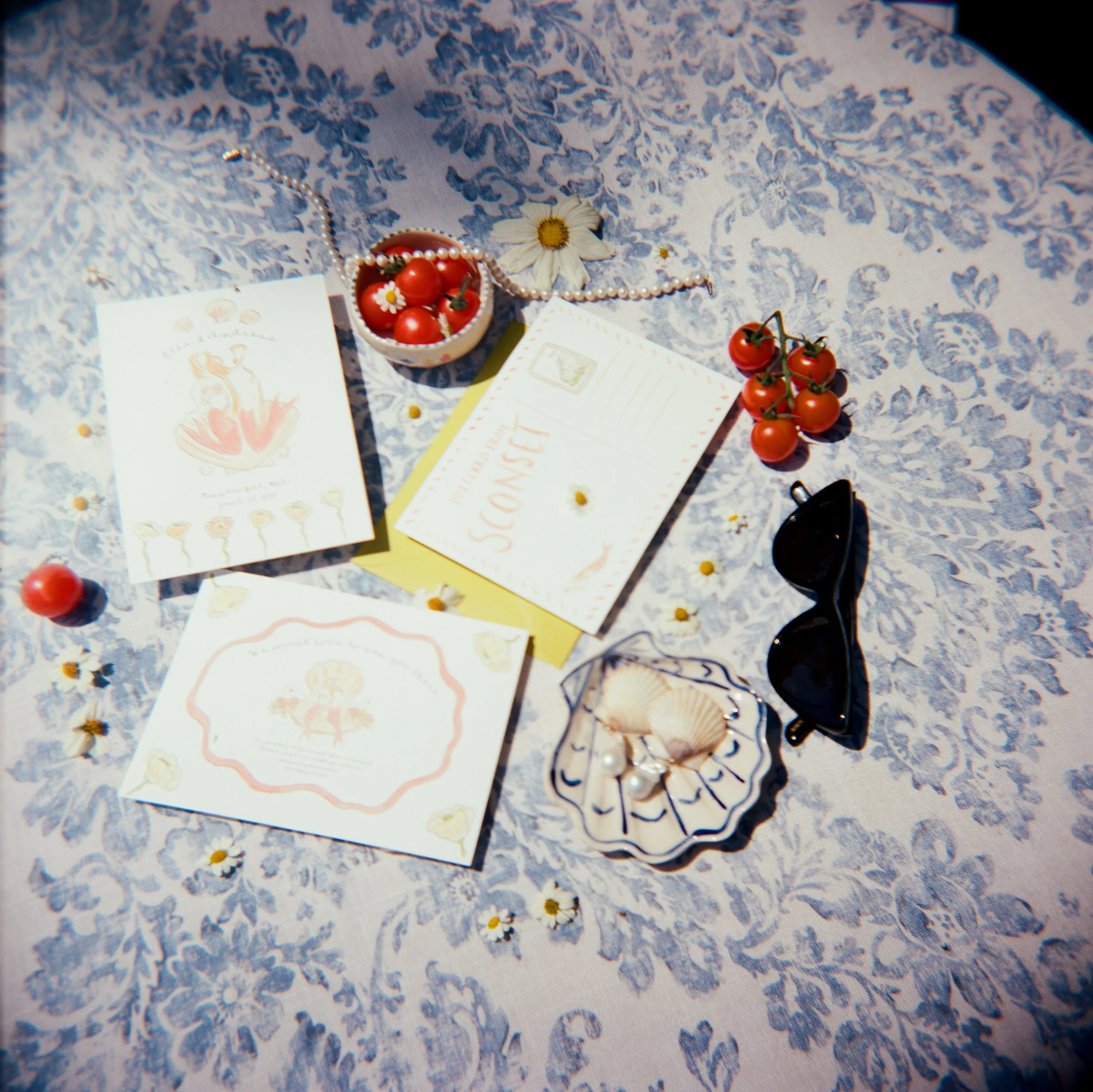 Summer-themed flat lay with cherry tomatoes, postcards, a seashell dish with pearls, a daisy flower, sunglasses, and scattered small daisies on a patterned cloth.