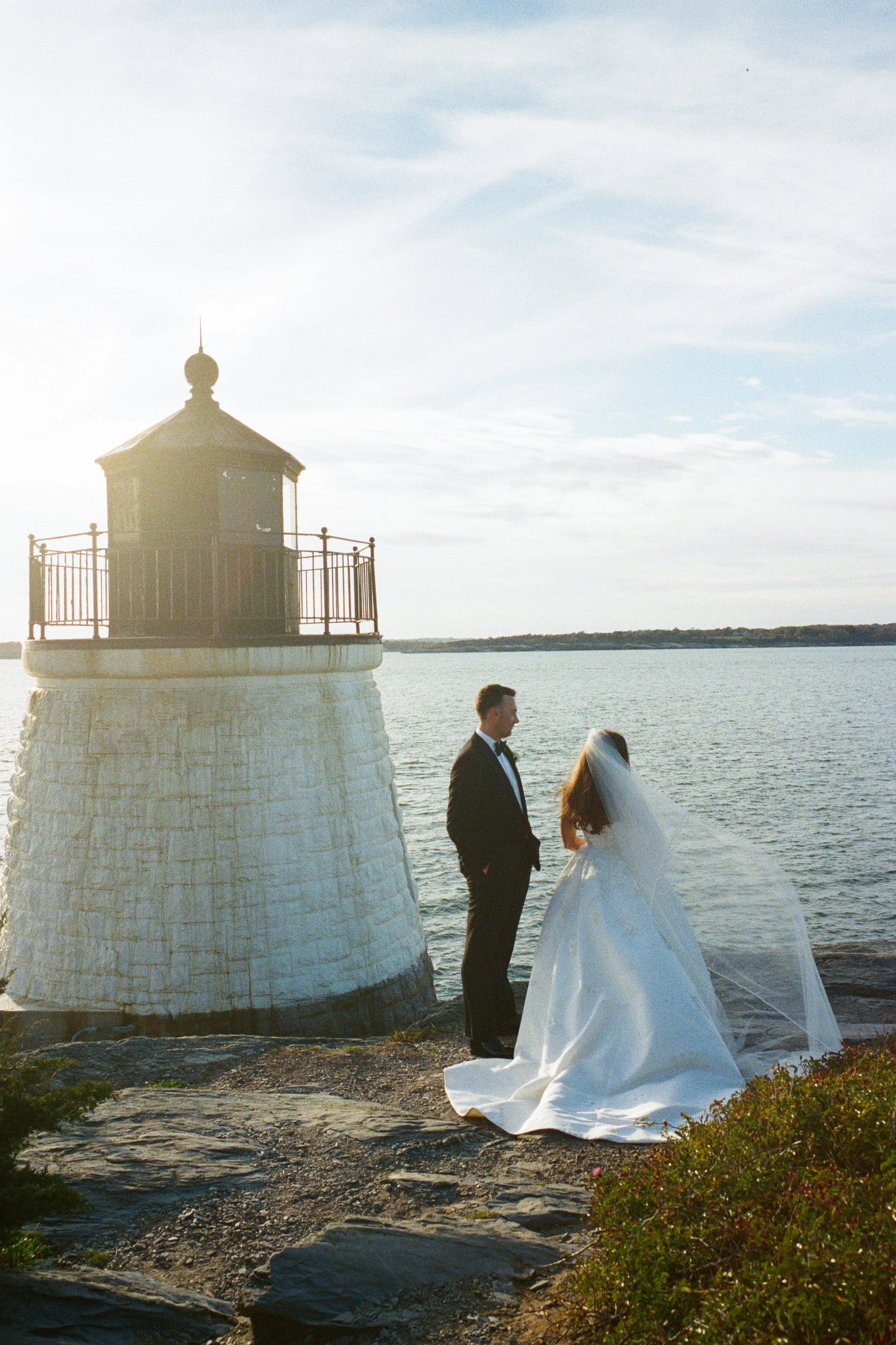 A bride and groom stand by a lighthouse near the water, with the bride wearing a white wedding gown and veil, and the groom in a black tuxedo, during sunset or sunrise.
