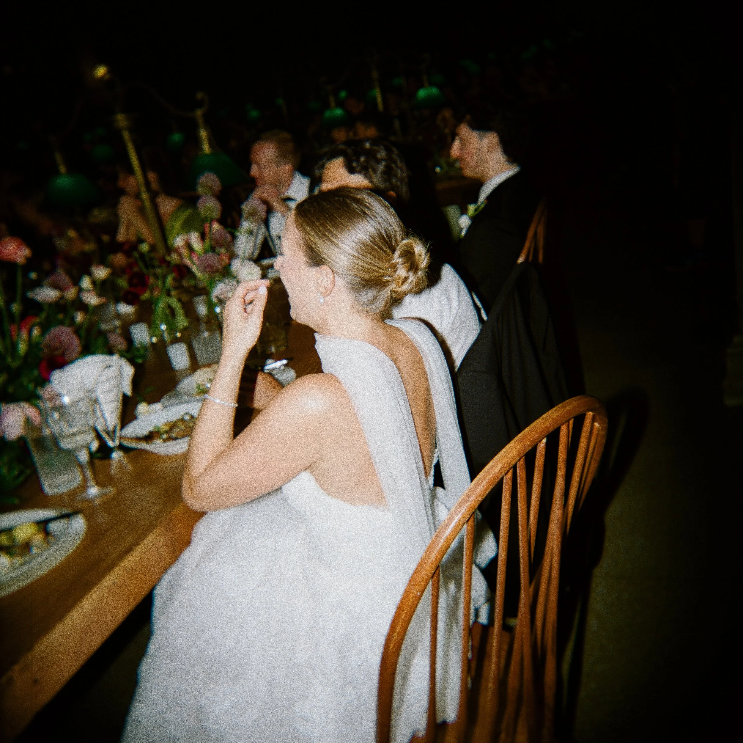 A bride in a white wedding dress with her hair in an elegant bun, sitting at a formal dinner table with other guests in suits and dresses, surrounded by floral centerpieces and dinnerware, at an indoor wedding reception.