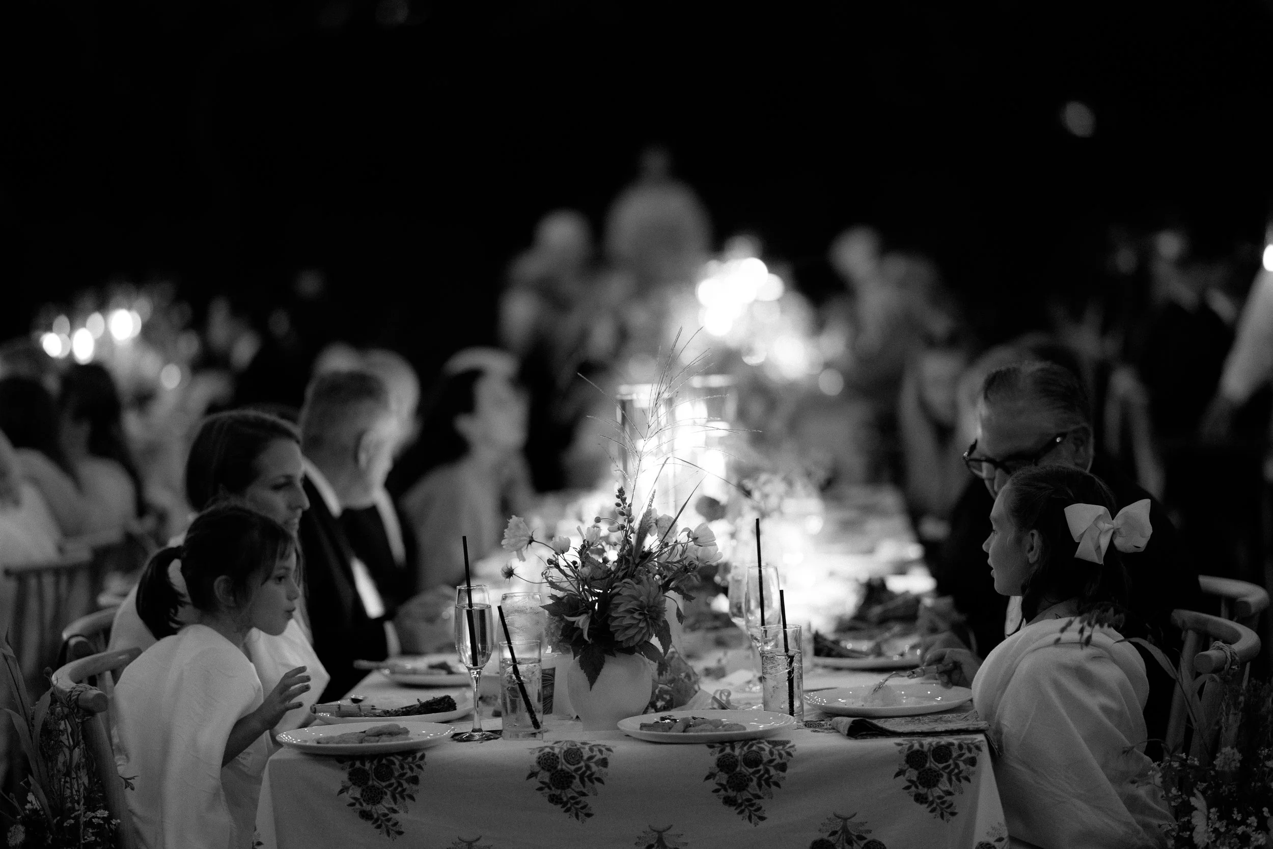 A black-and-white photo of a formal dinner party with multiple people sitting at a long table decorated with flowers. Some children and adults are visible, dressed in formal attire, enjoying the meal and conversation in a dimly lit setting.