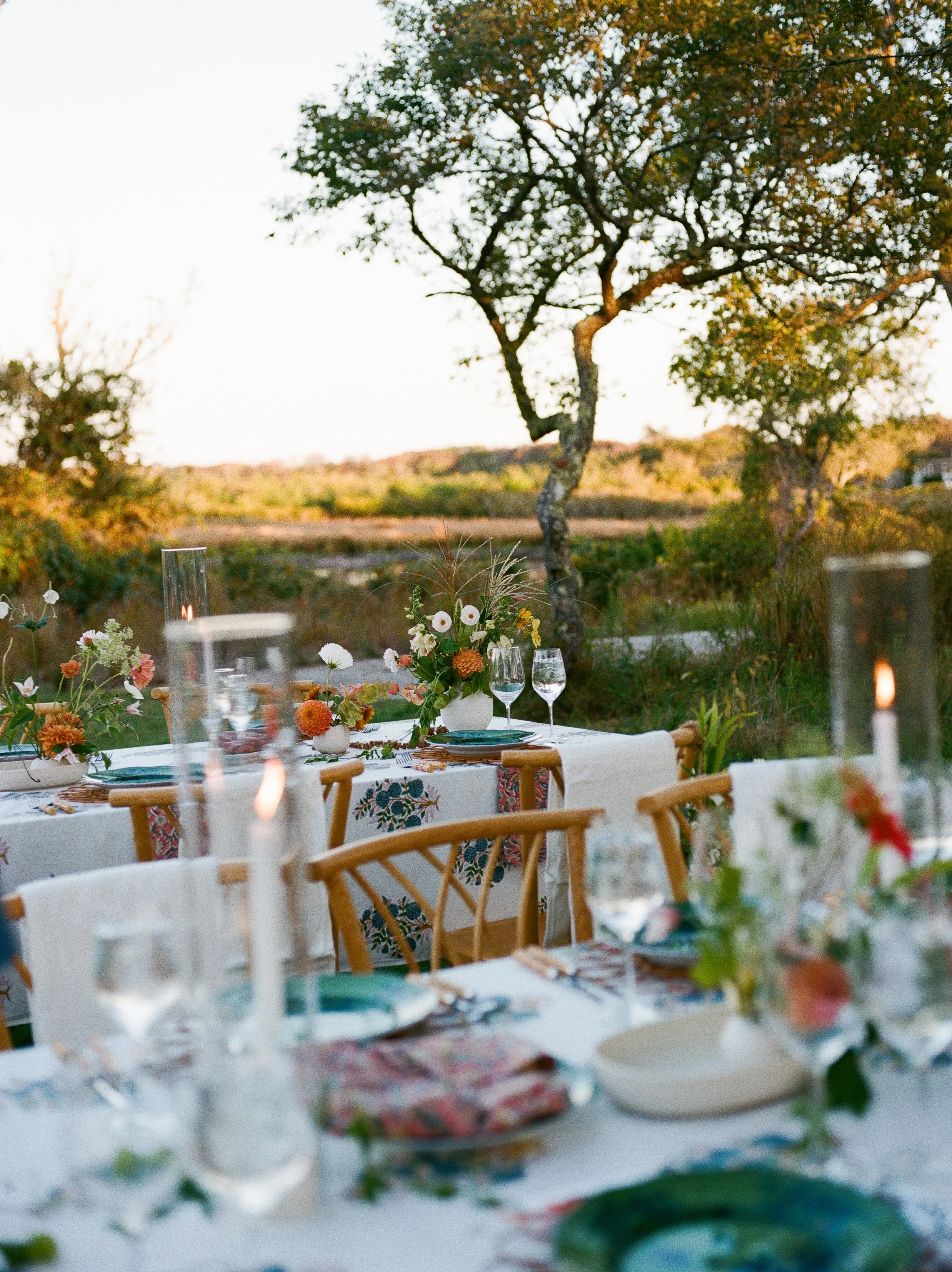 Outdoor dining table set for a meal with floral arrangements, candles, and glassware, along with chairs, overlooking a scenic landscape with trees and open land at sunset.