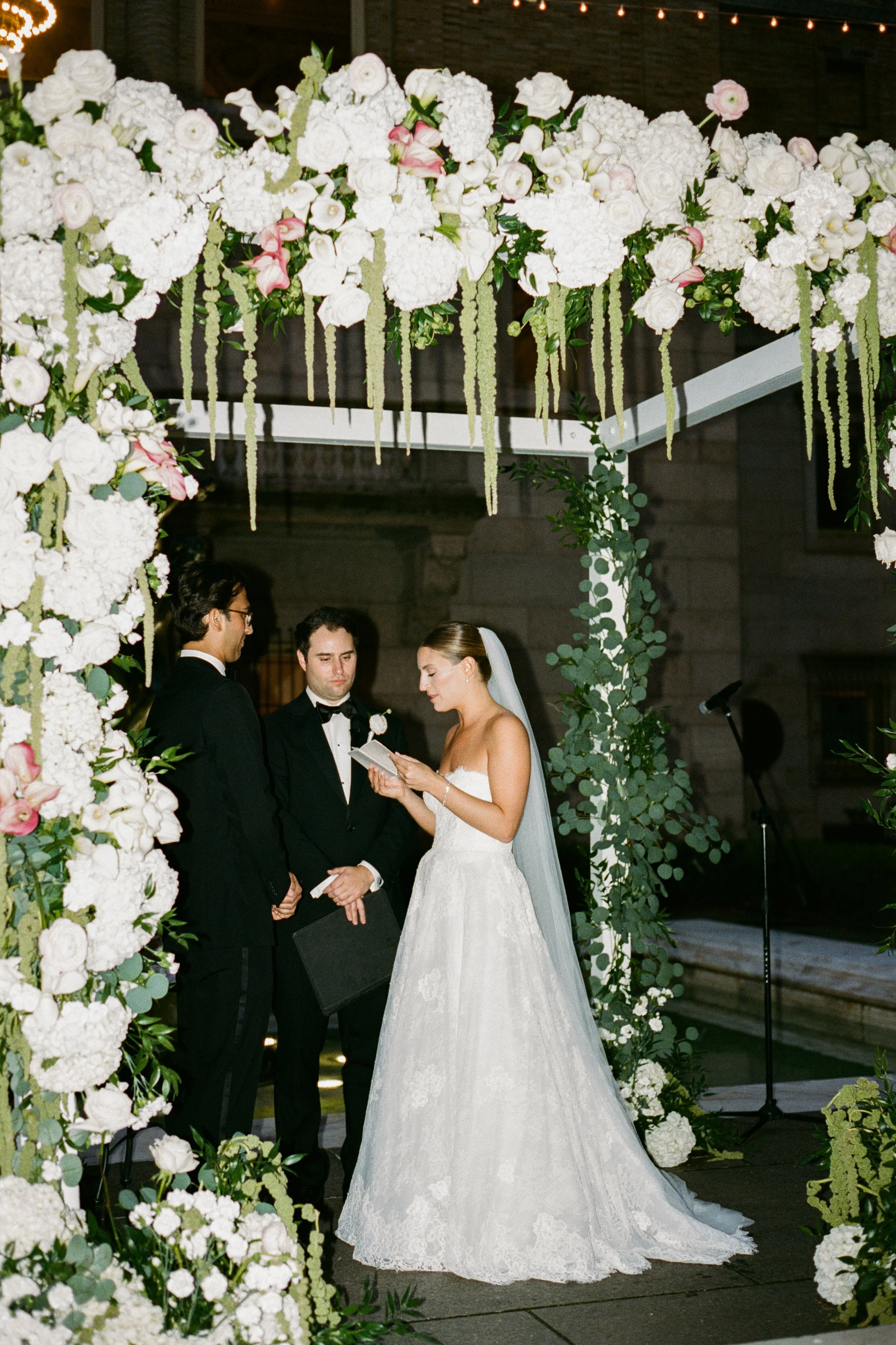 A bride reading vows during a wedding ceremony under a floral arch with white and pink flowers and greenery, at night.