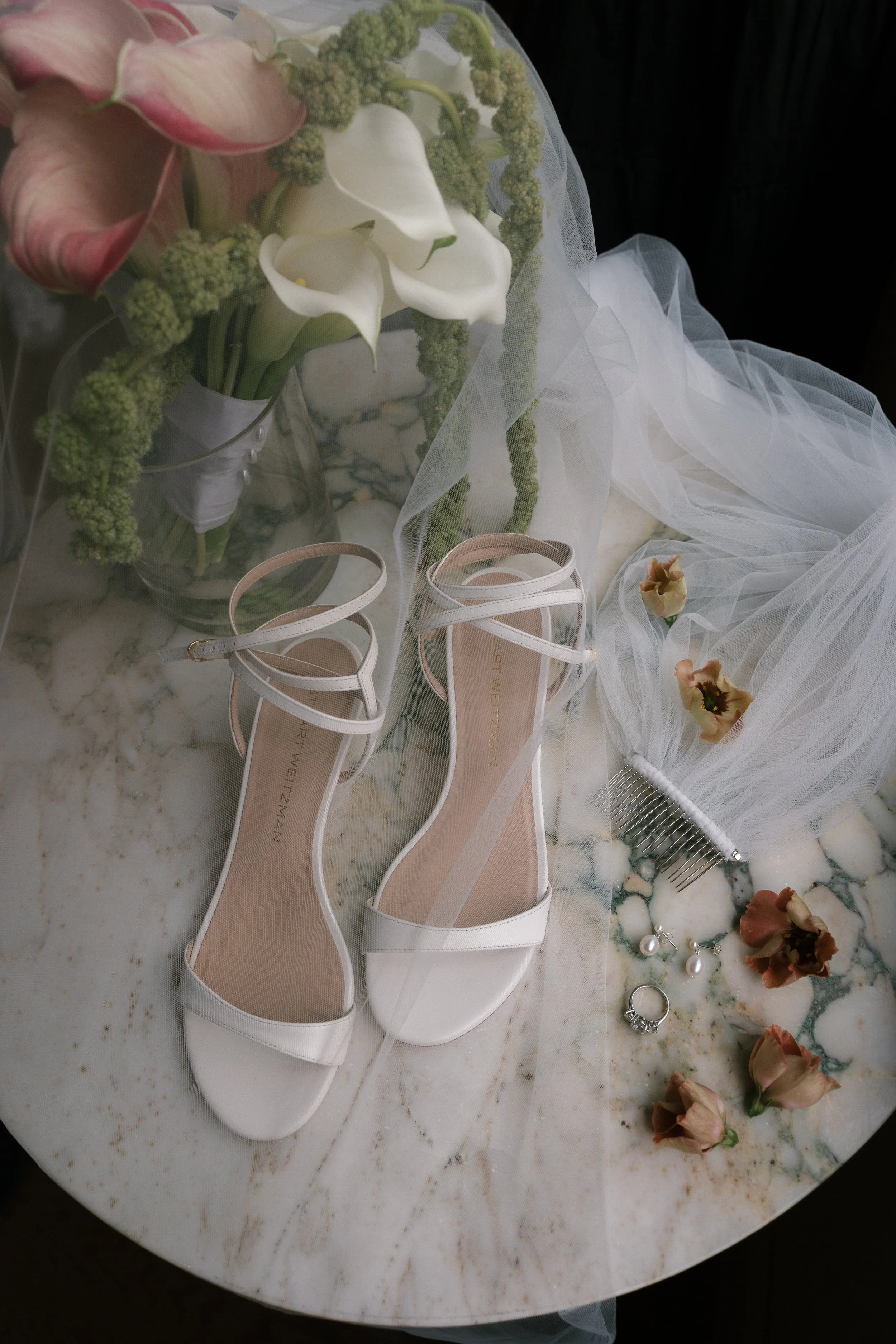 White high heel sandals with ankle straps, a wedding veil with dried flowers, pearl earrings, a ring, a pearl hair comb, a bouquet of pink and white flowers, and scattered dried roses on marble surface.