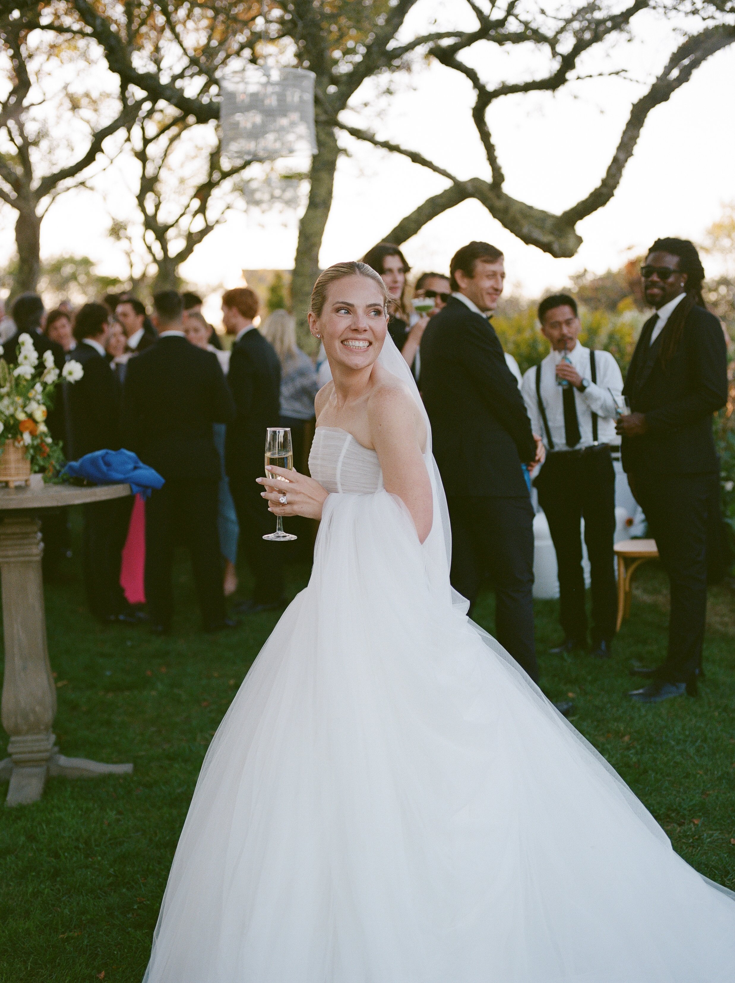 A bride in a strapless wedding gown with a full skirt smiling and holding a glass of champagne at an outdoor wedding reception, with guests mingling and trees in the background.