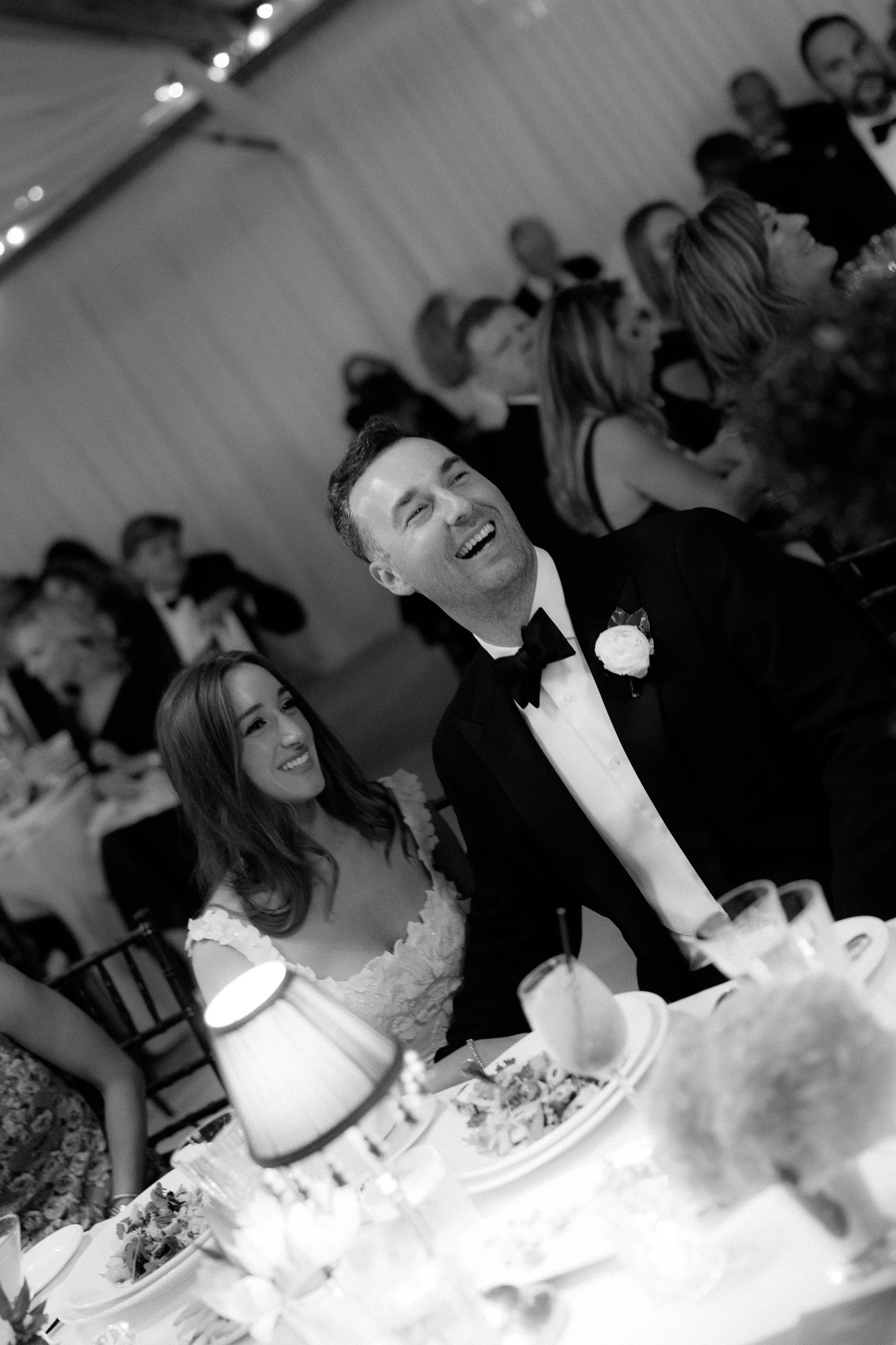 Black and white photo of a wedding reception with a laughing groom and a smiling bride seated at a table.