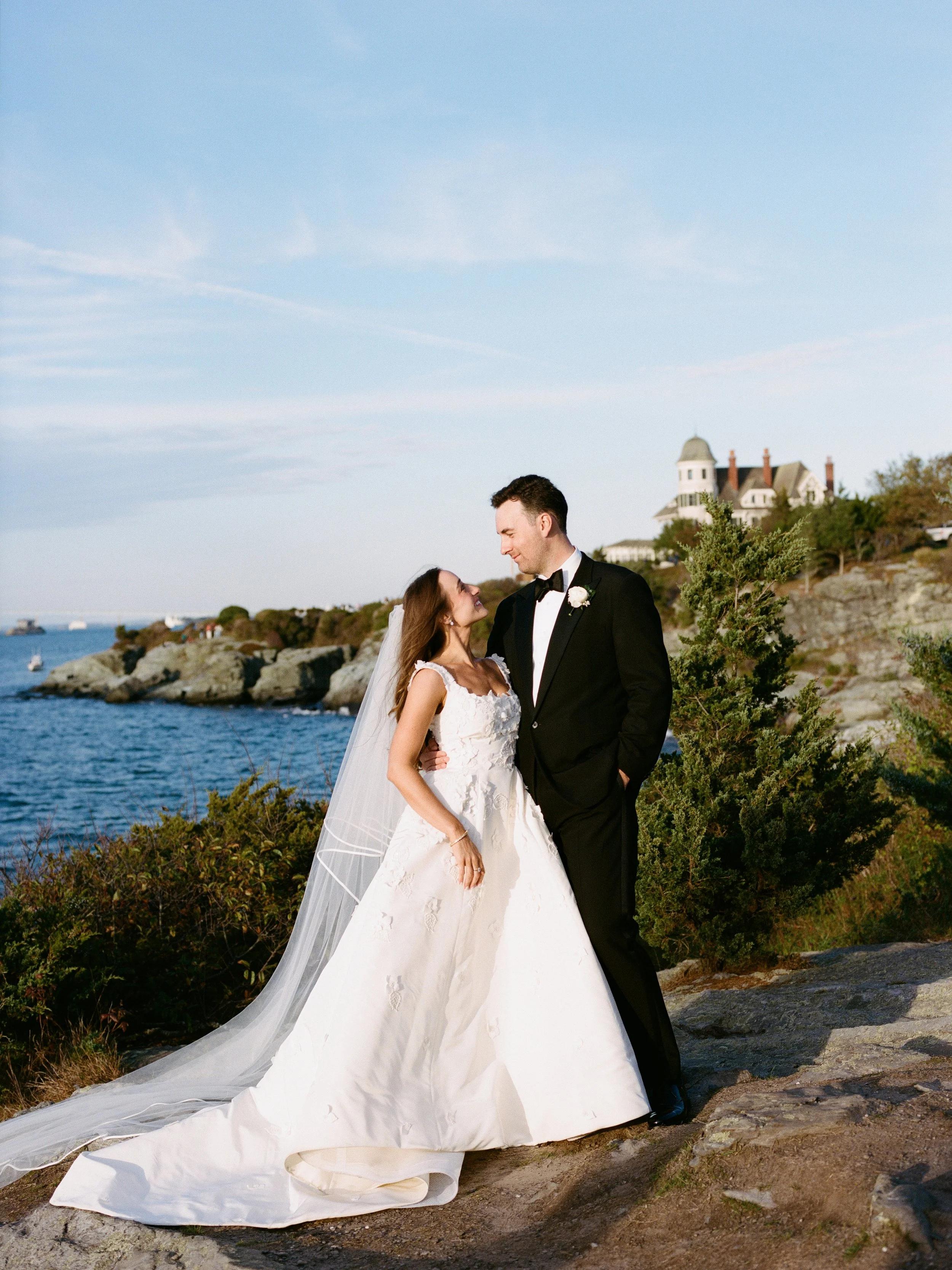A bride and groom standing together outdoors near the water, with a scenic rocky shoreline and a historic building in the background, on a clear day.