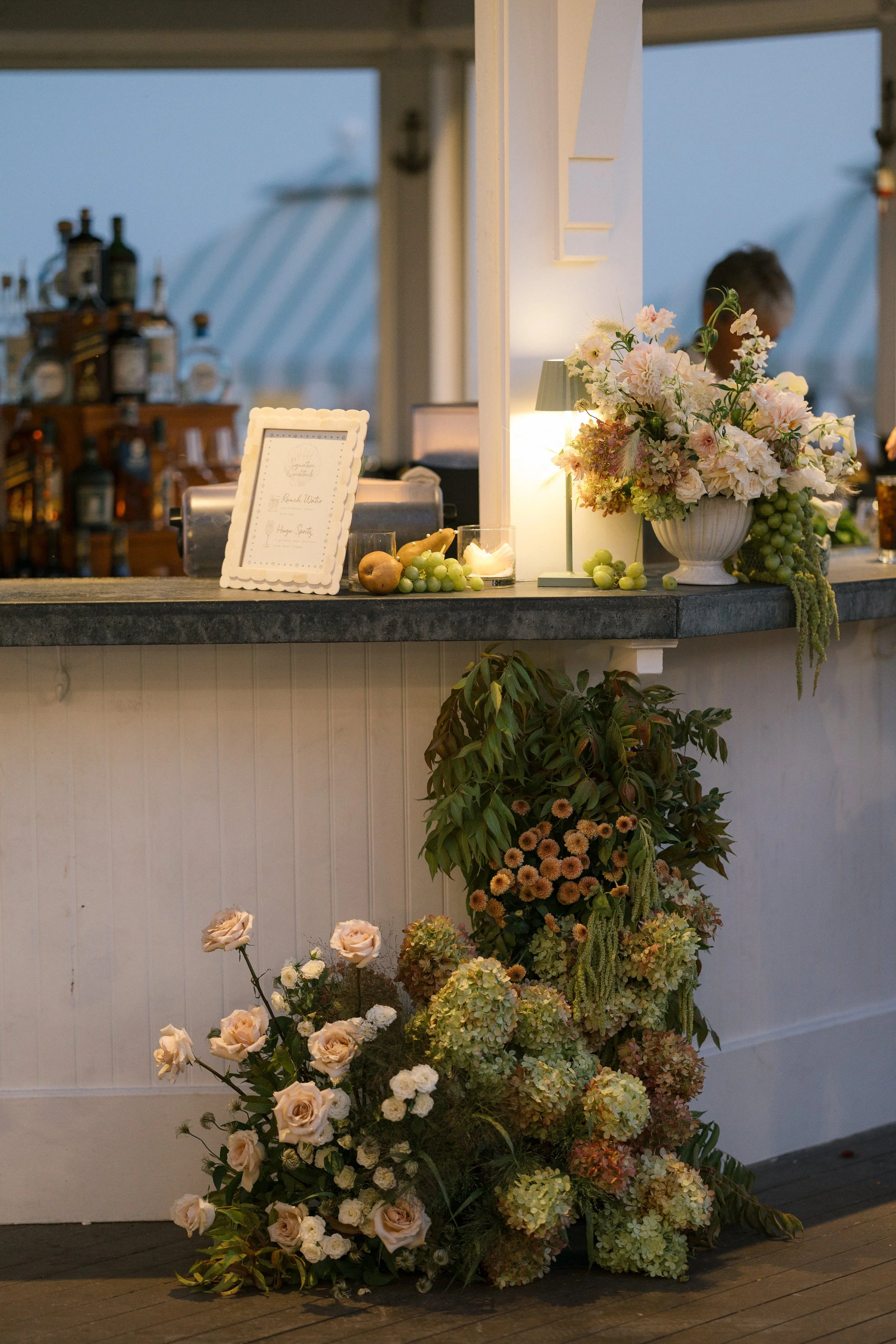A floral display on a bar counter includes a large white vase filled with pink and white flowers, a smaller candle, and a arrangement of green grapes and fruit. Below, on the floor, are various pink and cream flowers and green foliage, creating a lus