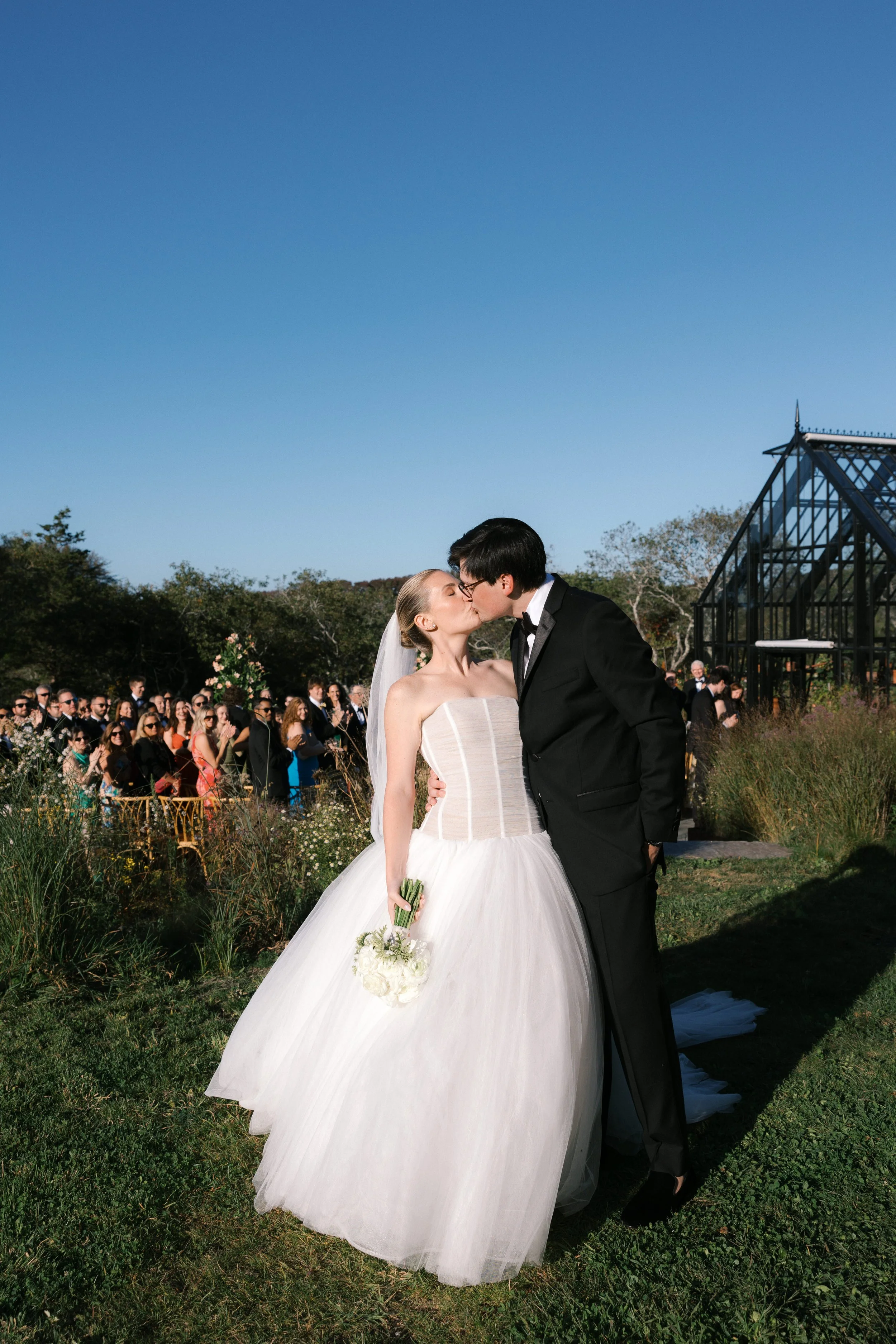 Bride and groom kissing at their outdoor wedding ceremony with guests in the background and a glass greenhouse structure to the right.