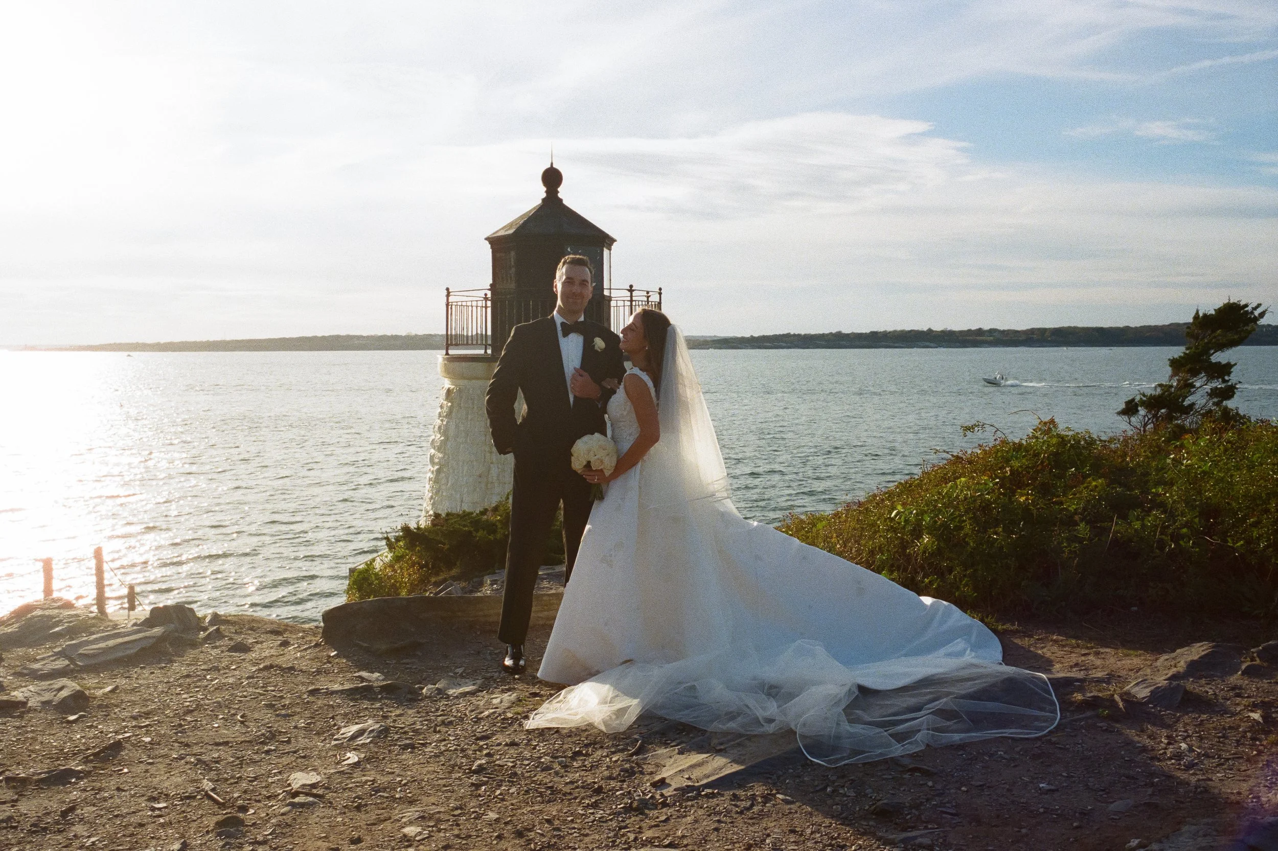 A groom in a black tuxedo and a bride in a white wedding gown standing together on a rocky shoreline by the water, with a small lighthouse in the background. The bride holds a bouquet of white flowers, and they are looking at each other during sunset