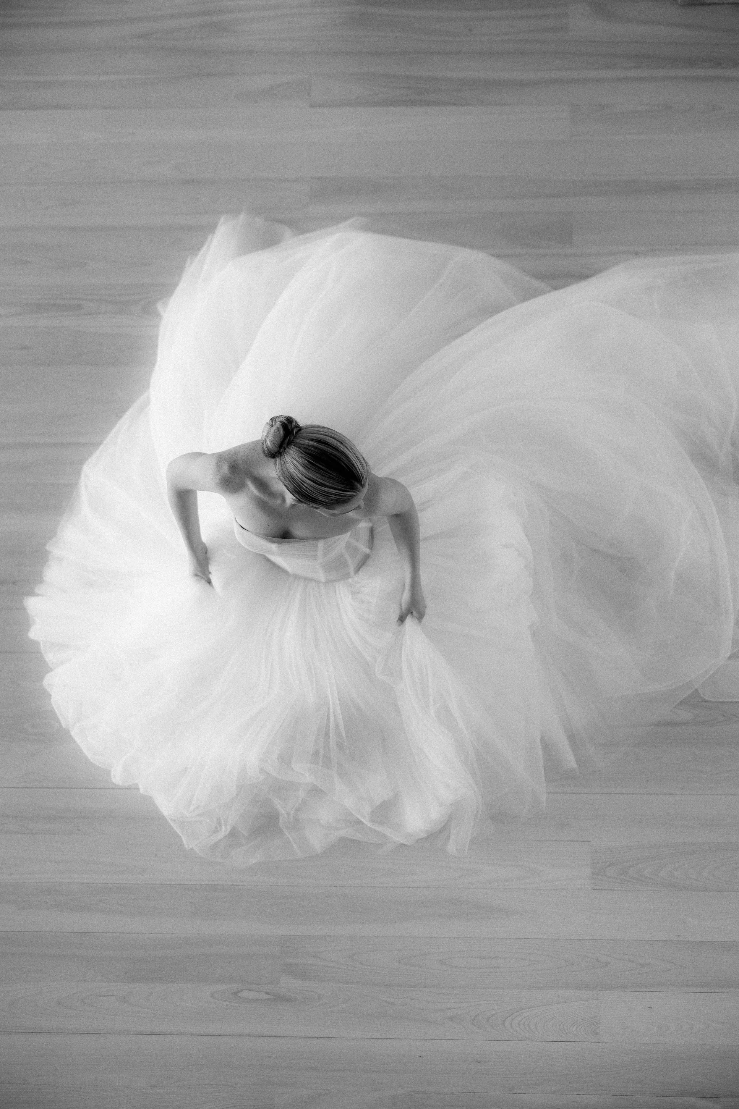 Black and white photograph of a woman in a flowing, strapless gown with a full skirt, viewed from above, standing on a wooden floor.