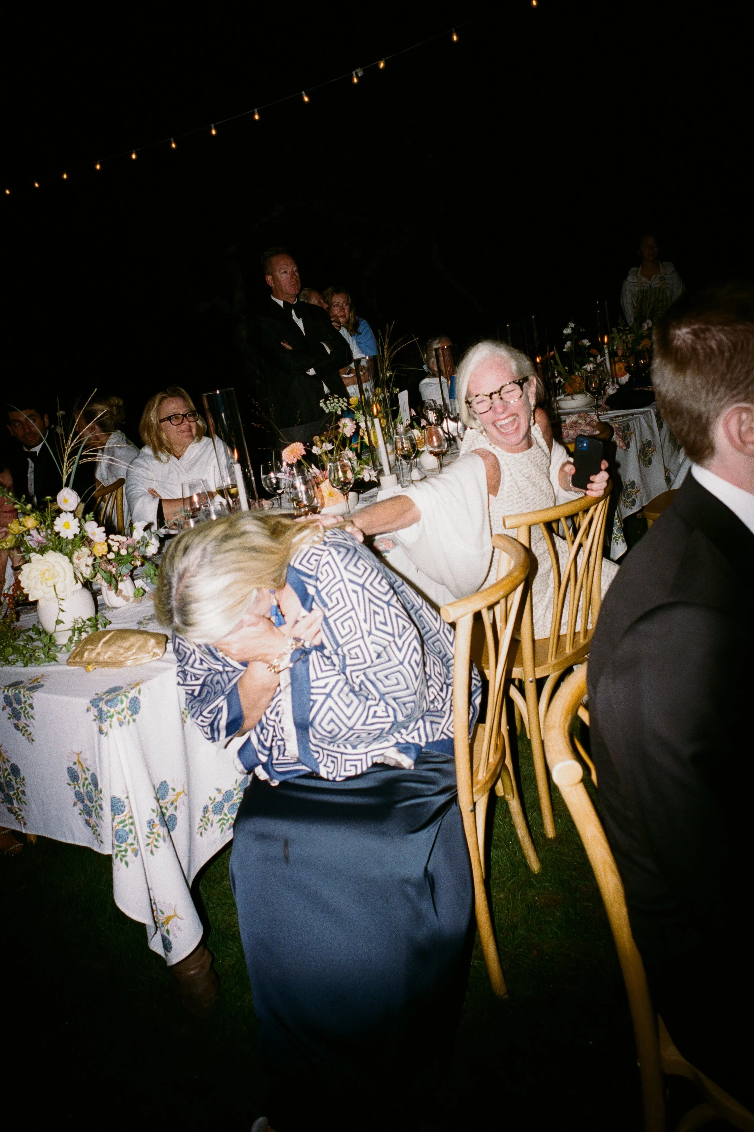 An outdoor nighttime celebration with elegantly set tables, floral centerpieces, and string lights overhead. People are dressed formally, with some standing and others seated, laughing and interacting.