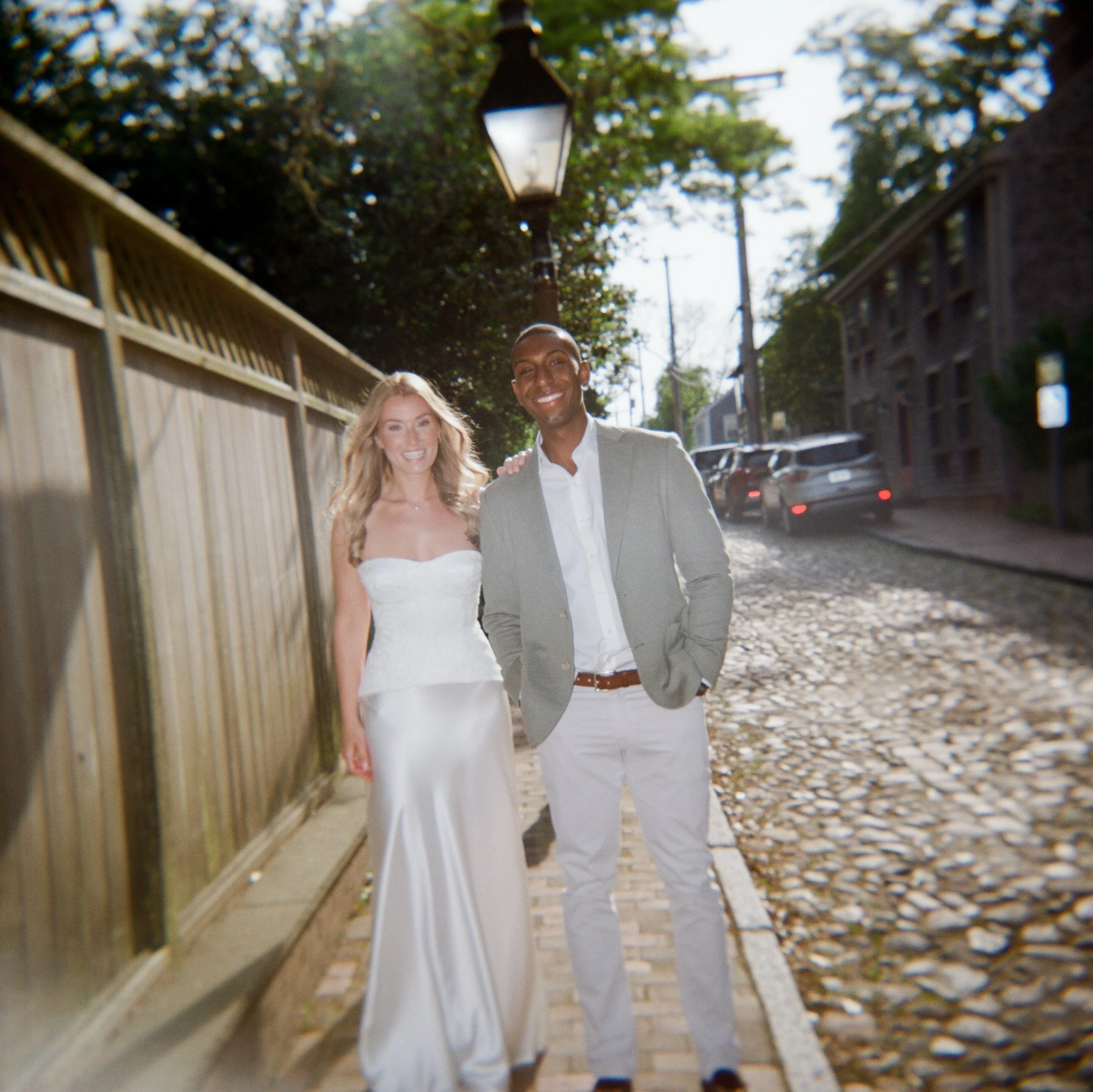 A smiling bride and groom walking arm in arm on a cobblestone street during daytime, with parked cars and trees in the background.