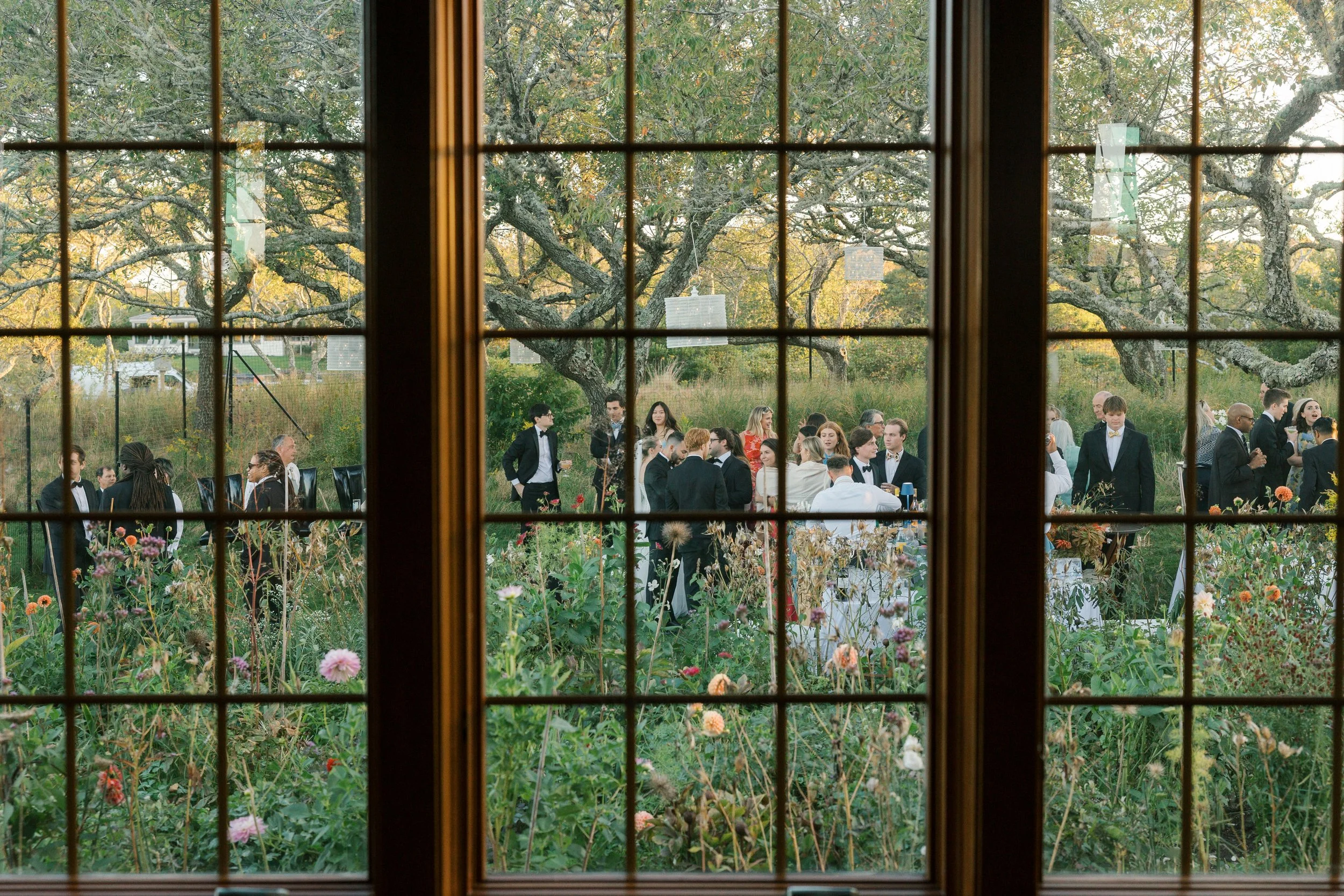 Gathering of people in formal attire outdoors viewed through a window with grid panes, with a garden and trees in the background.
