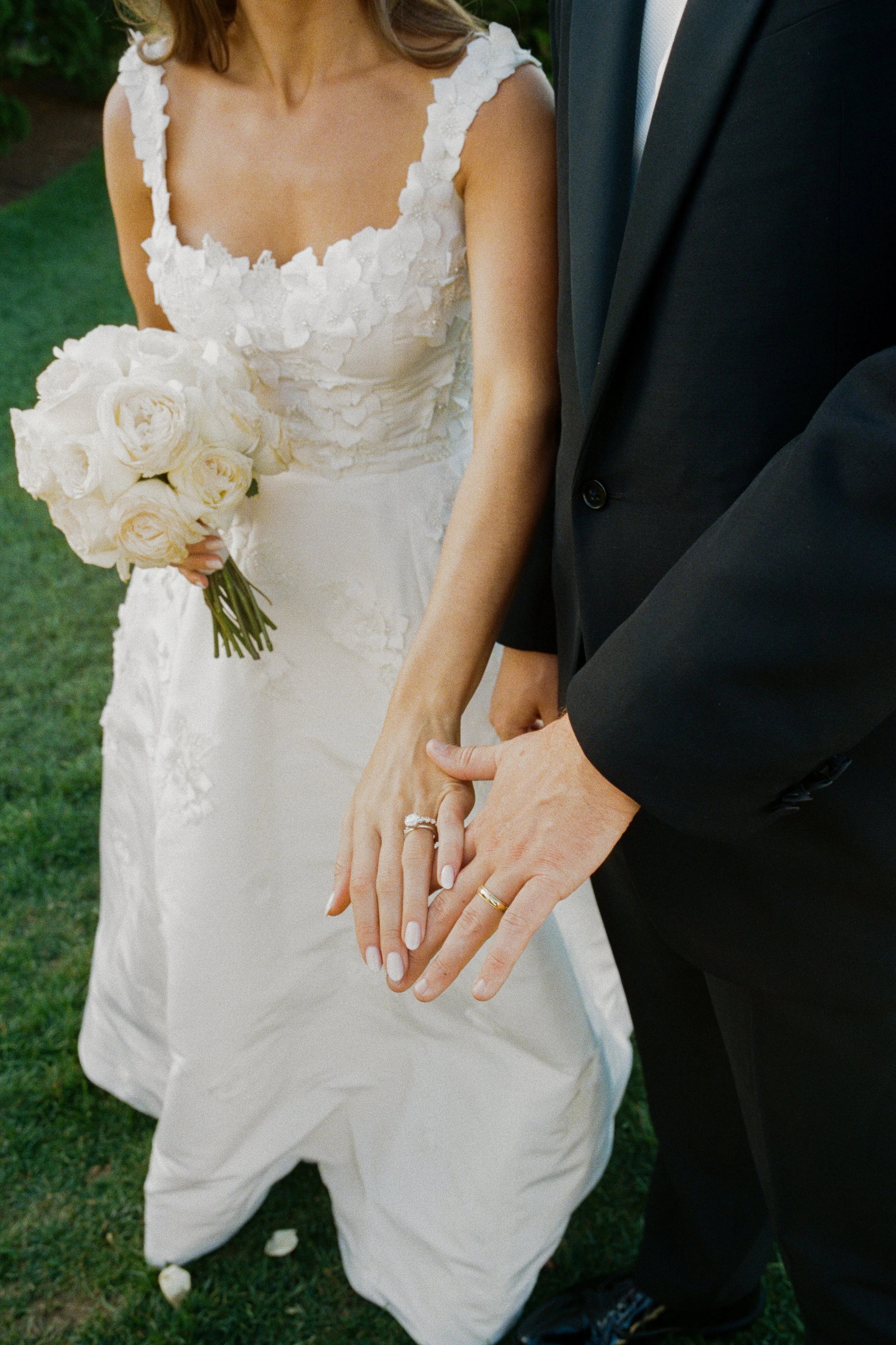 Close-up of a bride and groom holding hands, showing their wedding rings. The bride wears a white wedding dress and holds a bouquet of white roses, while the groom wears a black suit.