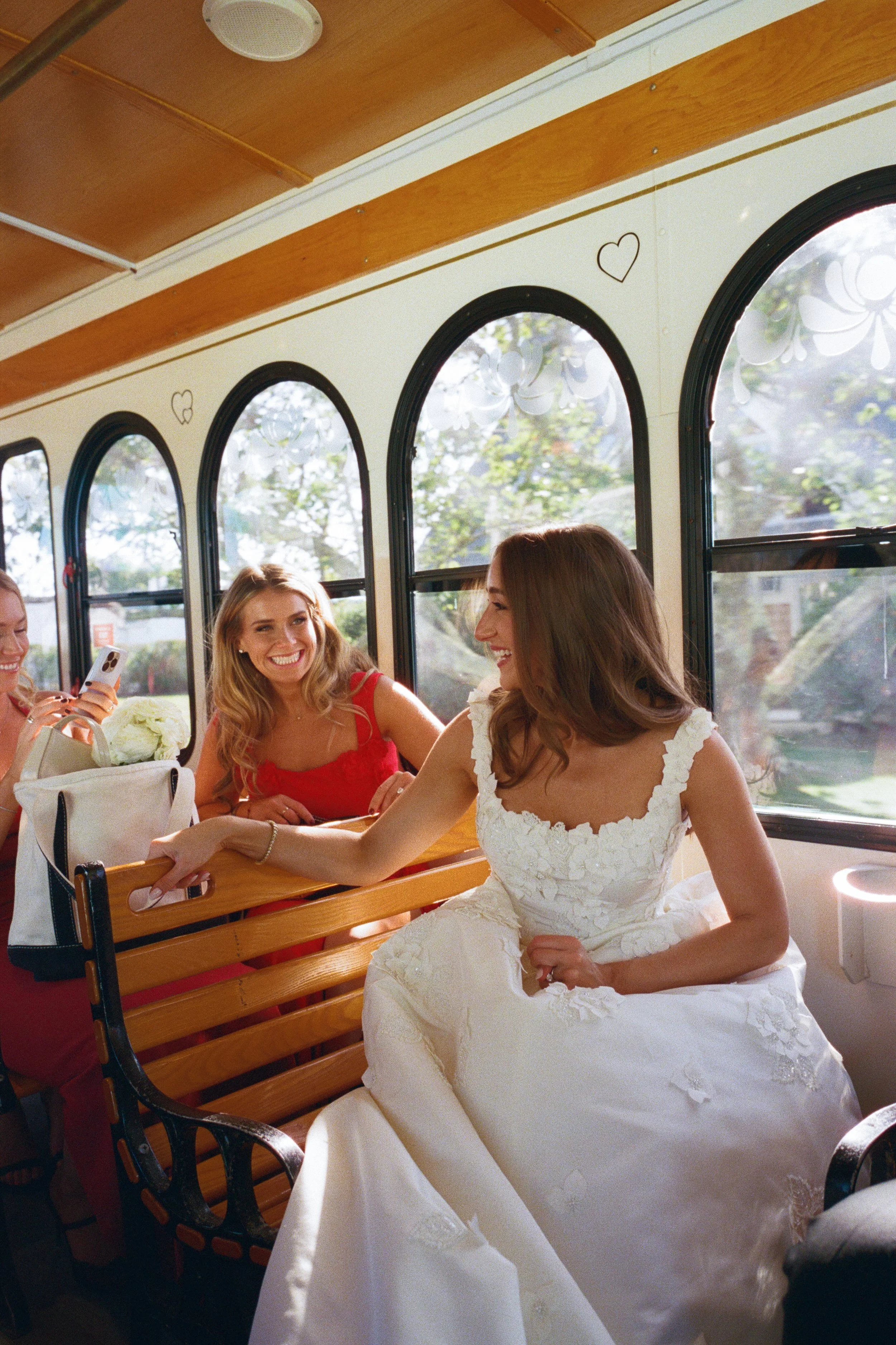 Women sitting on a wooden bench inside a trolley with arched windows, one in a wedding dress, smiling and engaging with others.