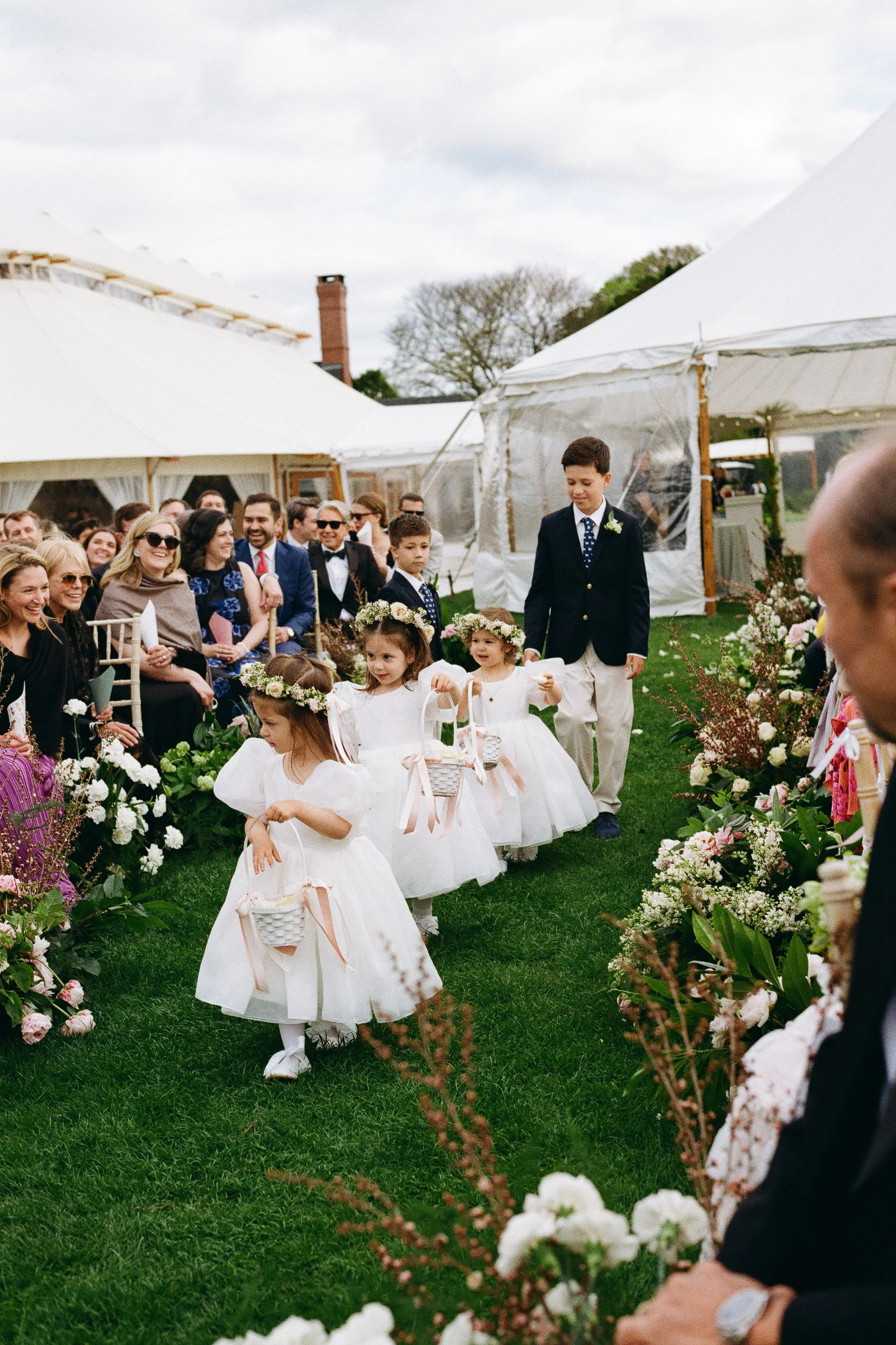 Young flower girls in white dresses and floral crowns walking down an outdoor aisle at a wedding, surrounded by guests and floral decorations.