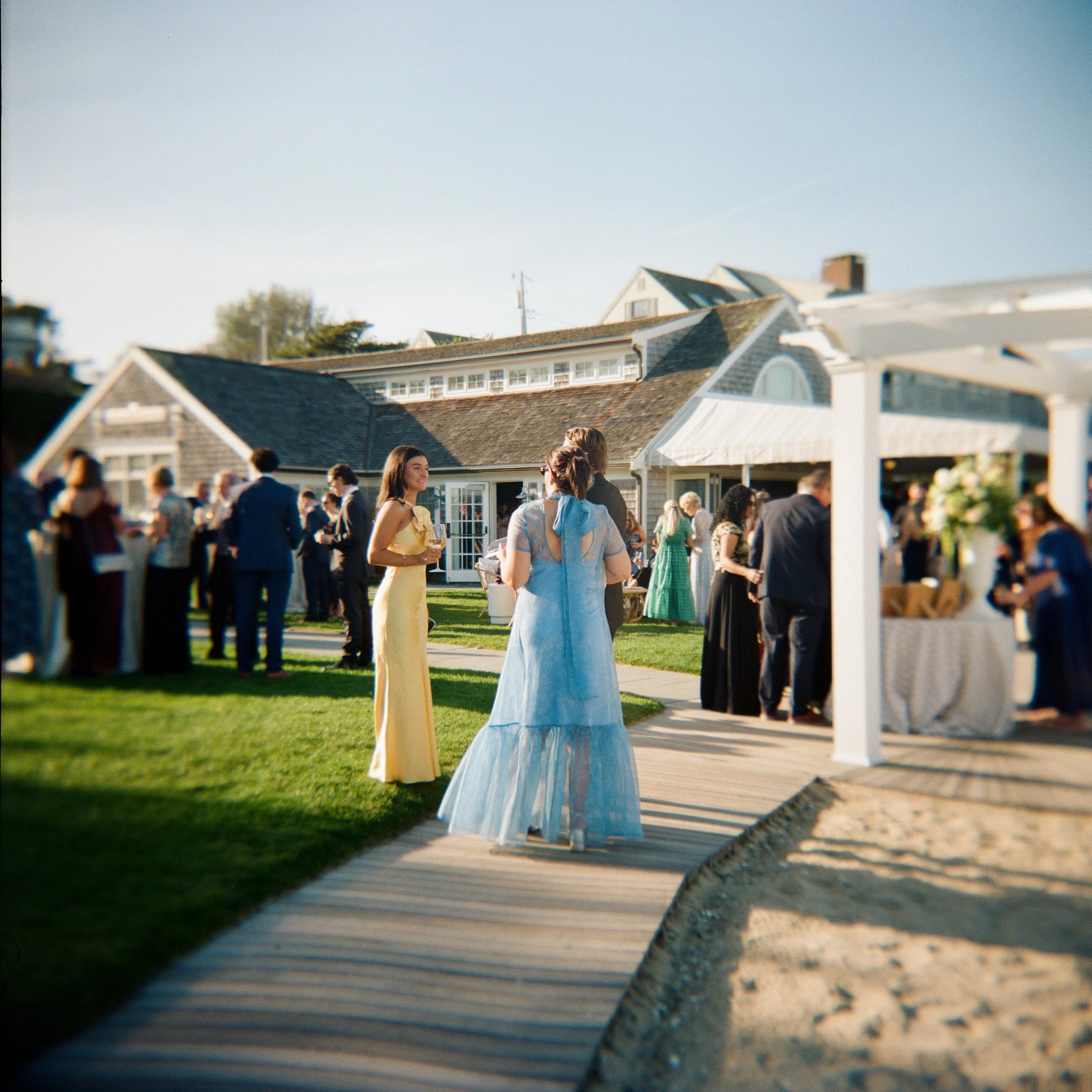 Guests gathered outdoors at a wedding reception, mingling on a grassy lawn near a house with a sloped roof and large windows on a sunny day.
