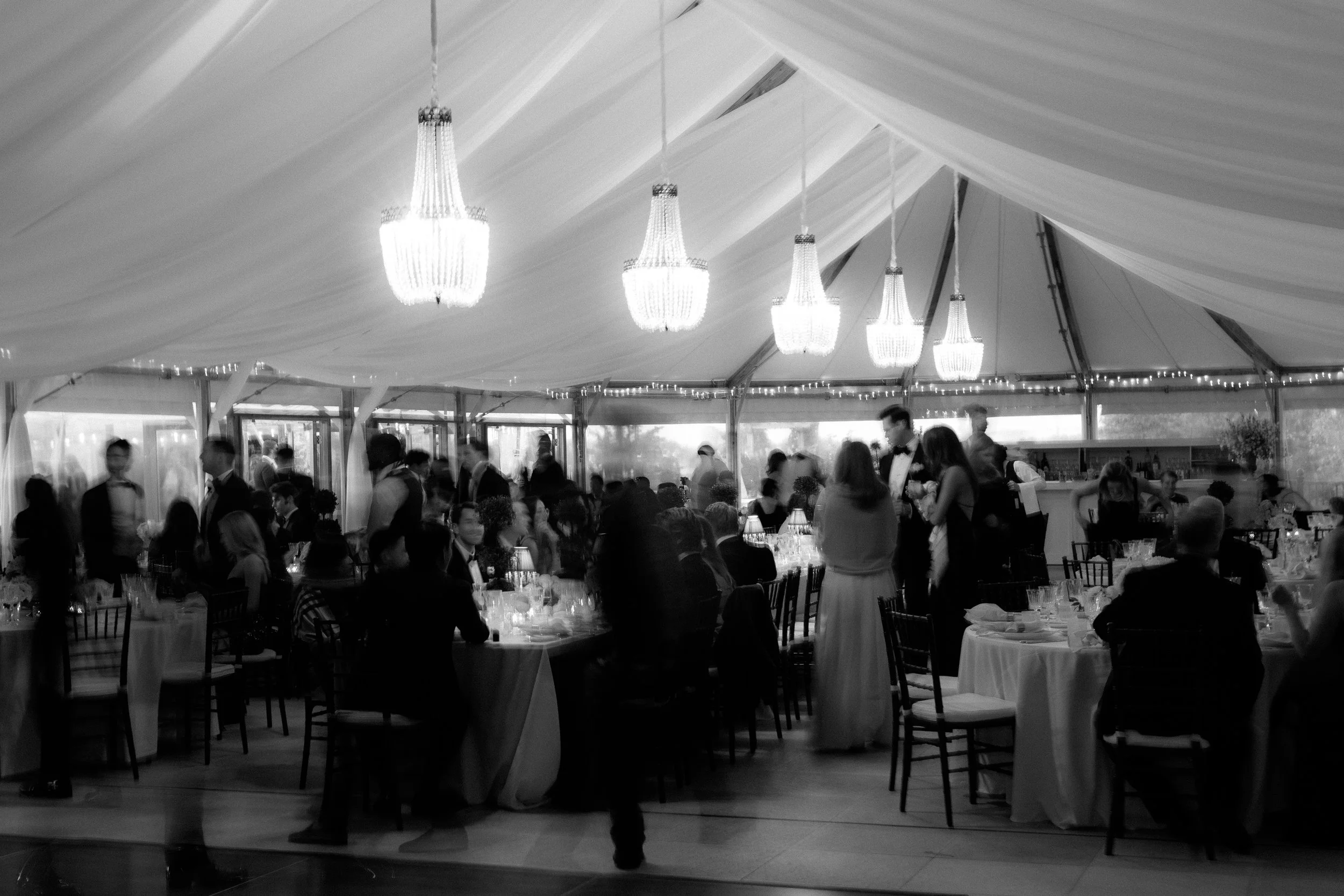 A black and white photo of a wedding reception in a decorated tent with chandeliers. People are seated at round tables and standing, with a bar in the background.